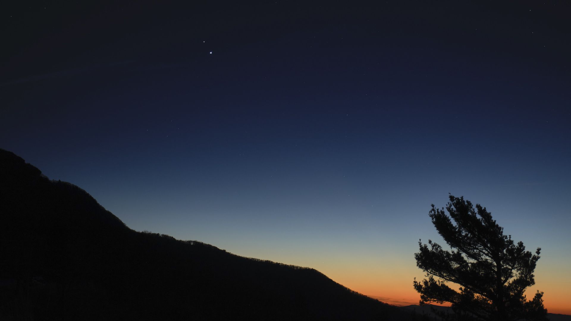Jupiter and Saturn seen shining like pinpoints of light, close together as the Sun sets behind a mountain and a gnarled tree
