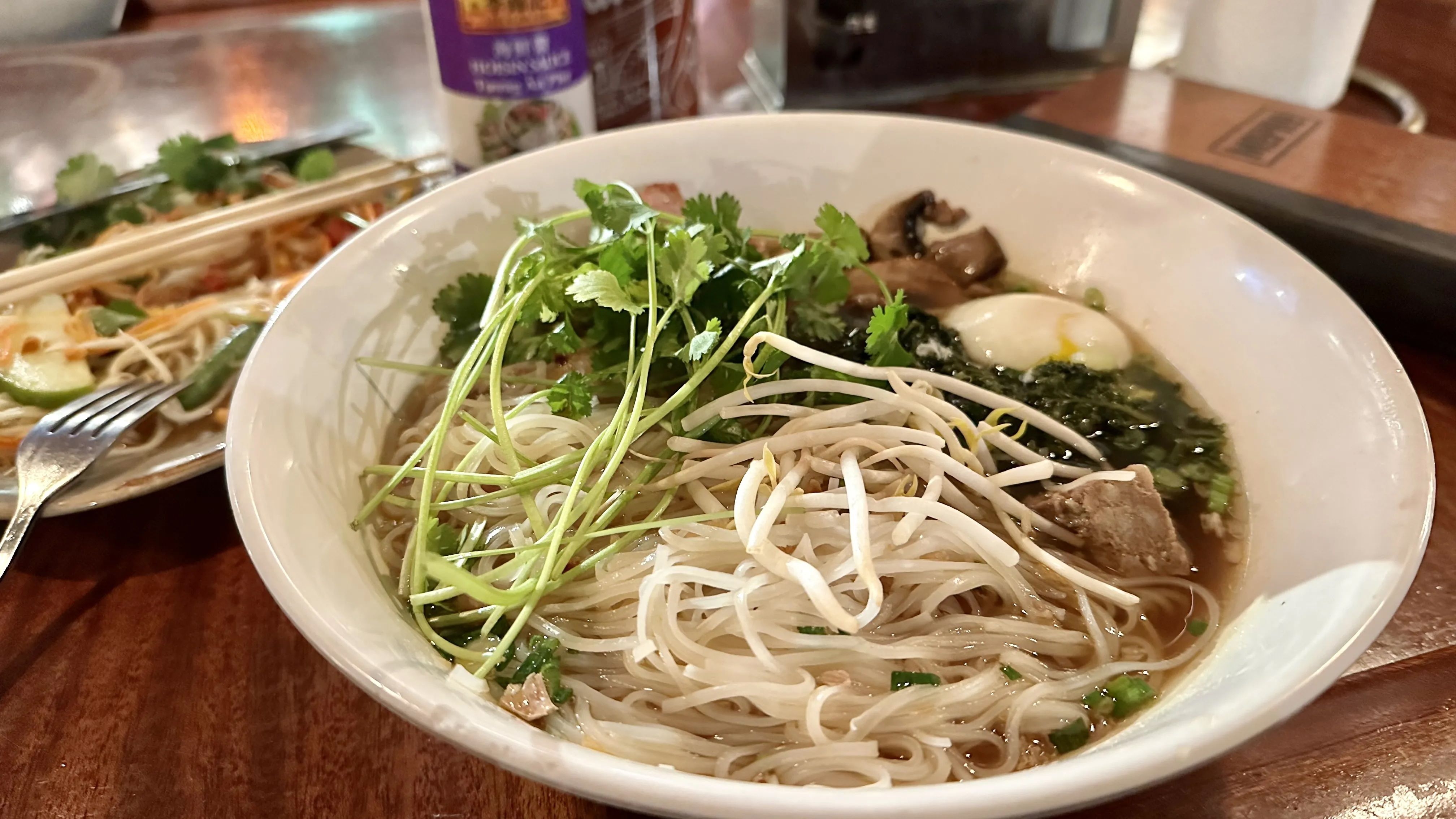 Bowl of noodle soup with cilantro, bean sprouts, mushrooms, boiled egg, and meat in broth on wooden table, with partially eaten dish and chopsticks in background.