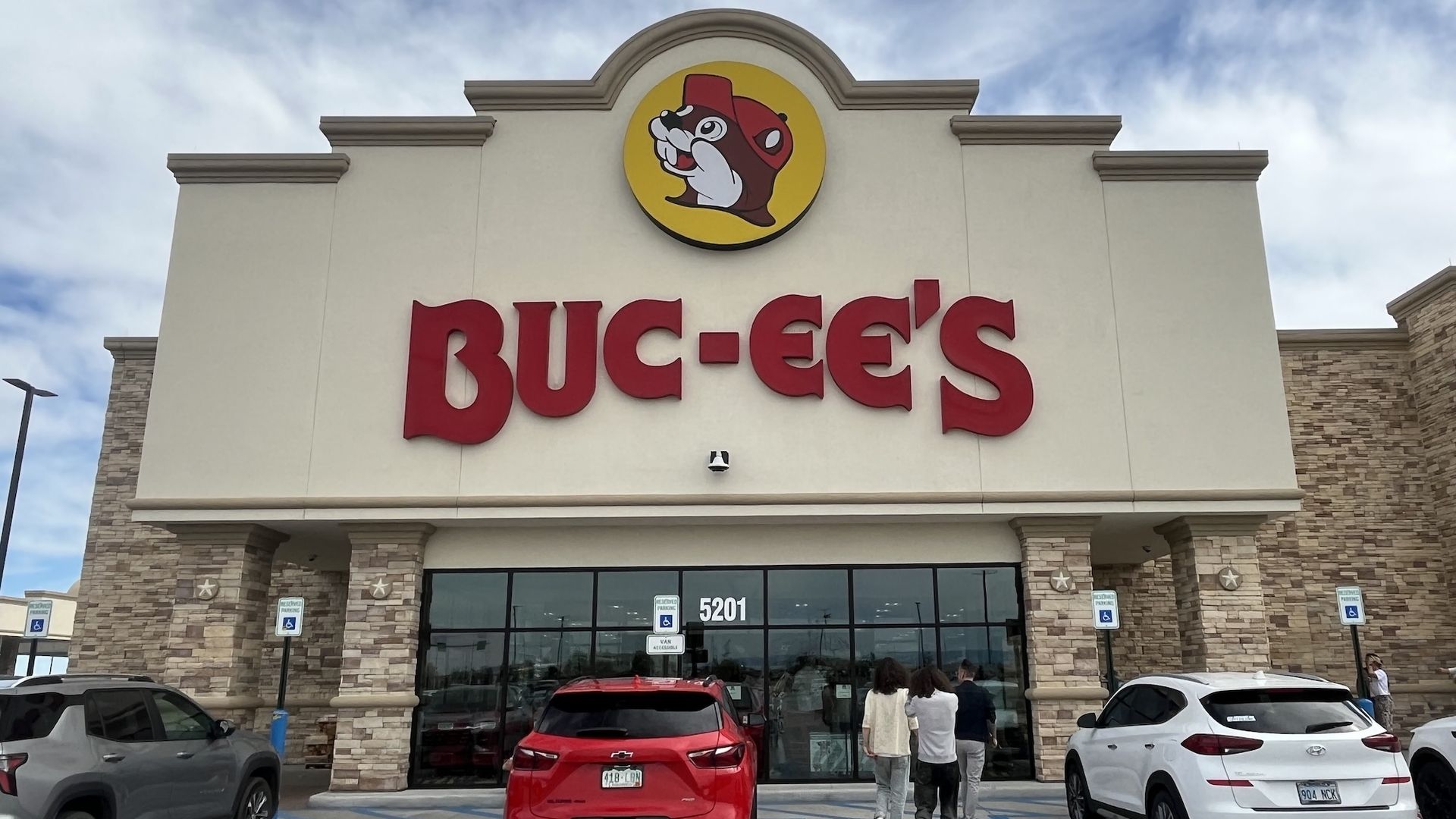 Front entrance of a Buc-ee's store with large red letters and beaver mascot logo above. Three people walk towards glass doors. Several parked cars and handicapped signs visible.