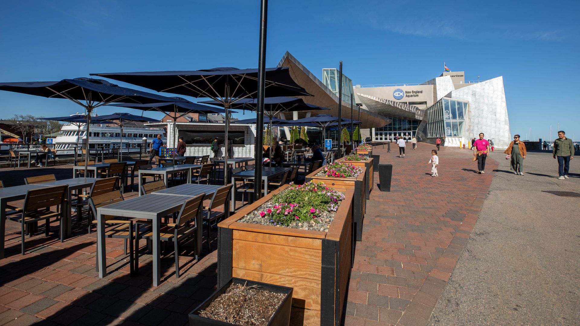 A picture of the Dockside beer garden, with open umbrellas and empty tables, being set up, with the New England Aquarium in the background.