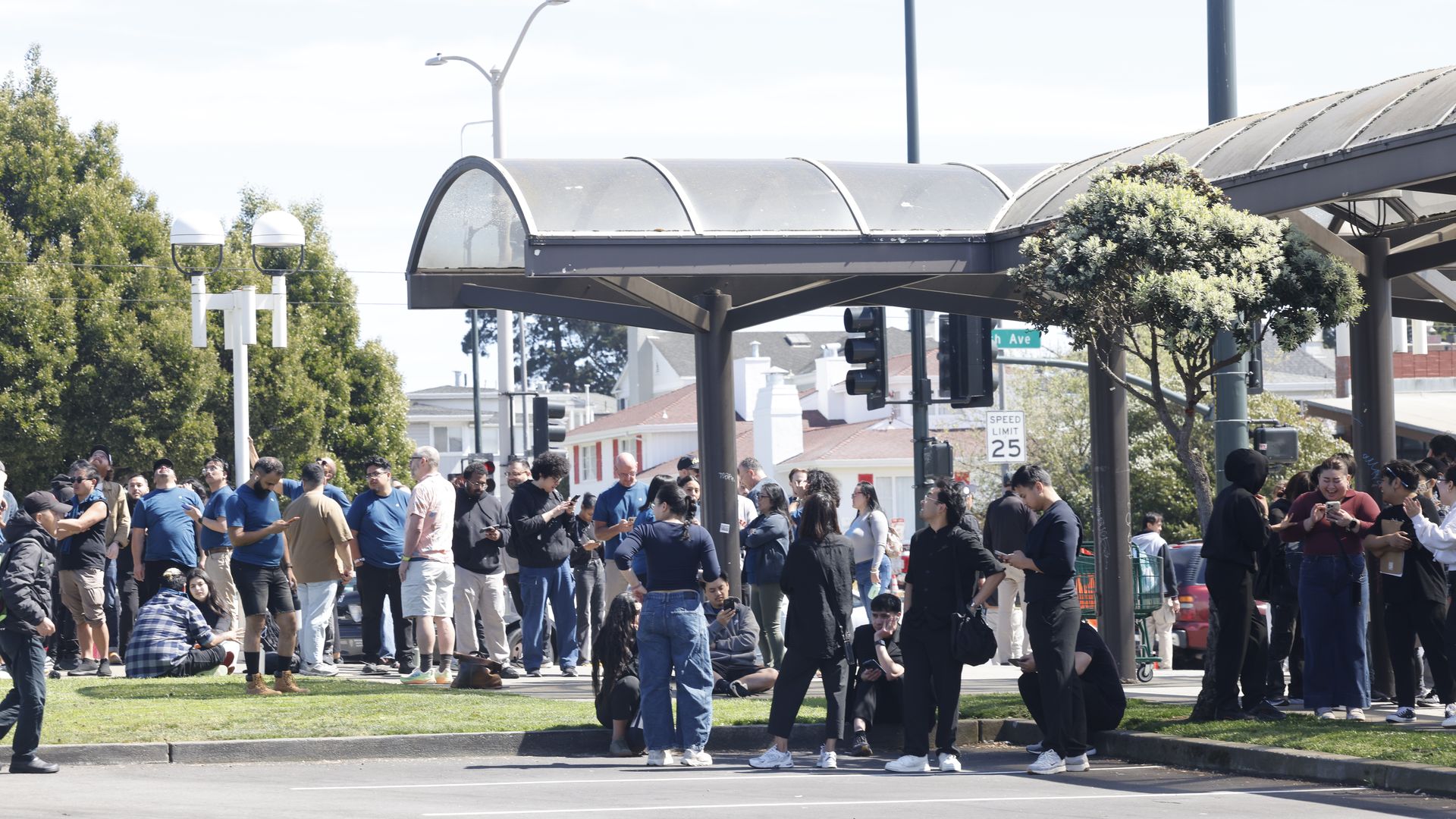 A large group of people waits along a sidewalk beneath a curved glass canopy in a sunny urban area, with street signs, traffic lights, trees, and cars in the background.