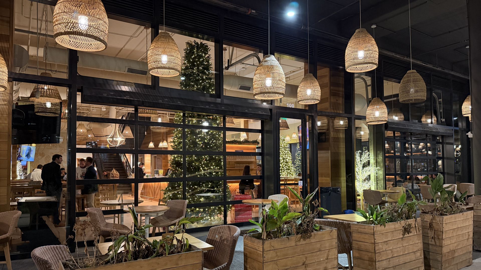 Outdoor and indoor seating at a restaurant decorated with large Christmas trees with white lights, wicker pendant lamps, wooden planters, and patrons inside at night.