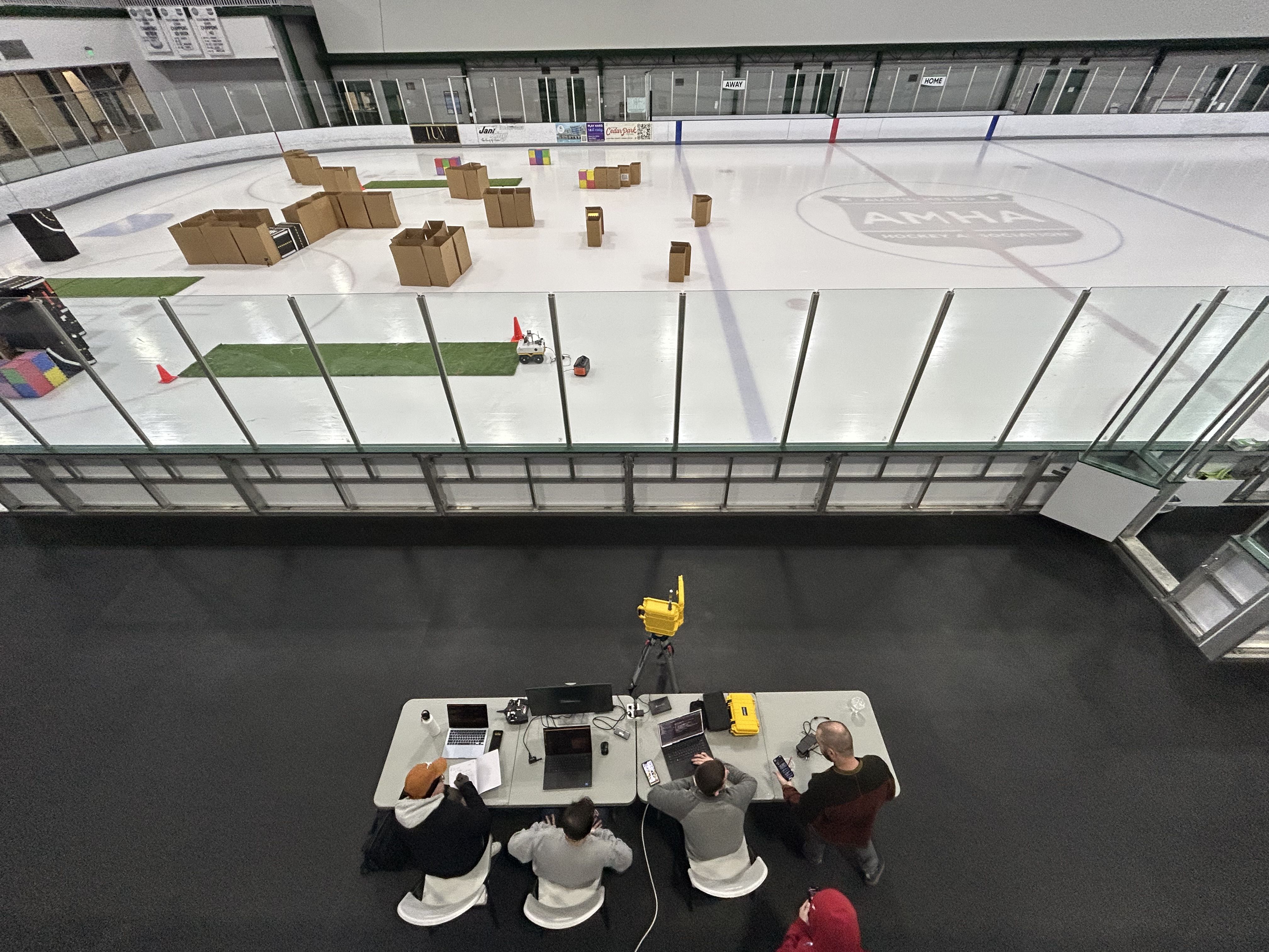 An indoor ice rink from above with scattered cardboard boxes forming a makeshift obstacle course; cardboard blocks, cones, and colored tiles on ice. Behind glass, people sit at a table with laptops.