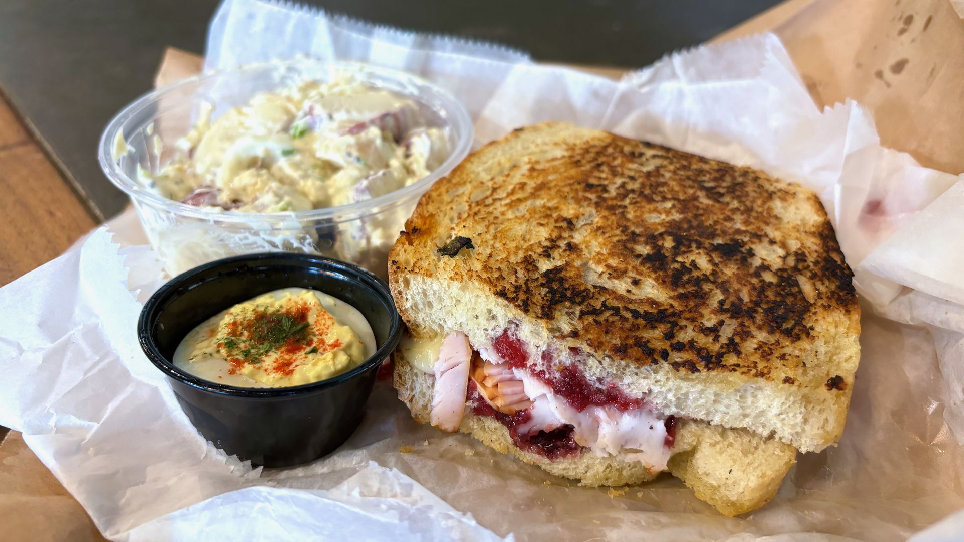 A large turkey sandwich on grilled sourdough bread next to a cup of potato salad and a deviled egg