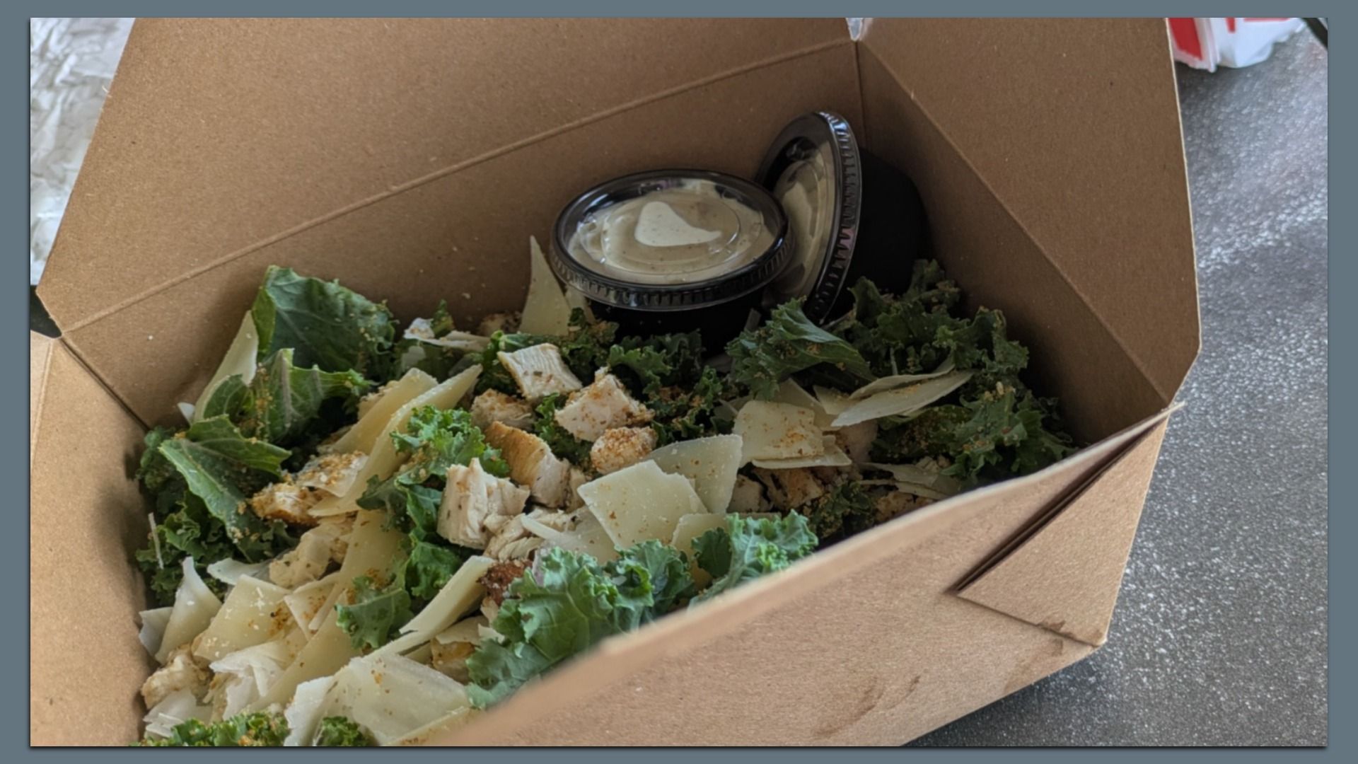 Takeout box with kale salad containing chopped chicken, shaved cheese, and two small containers of creamy dressing on the side.