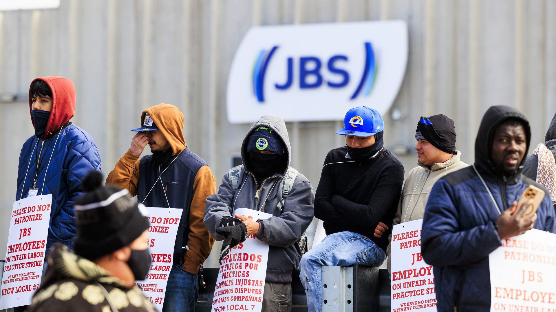 Striking workers stand outside a building with a JBS sign, holding white placards alleging unfair labor practices.
