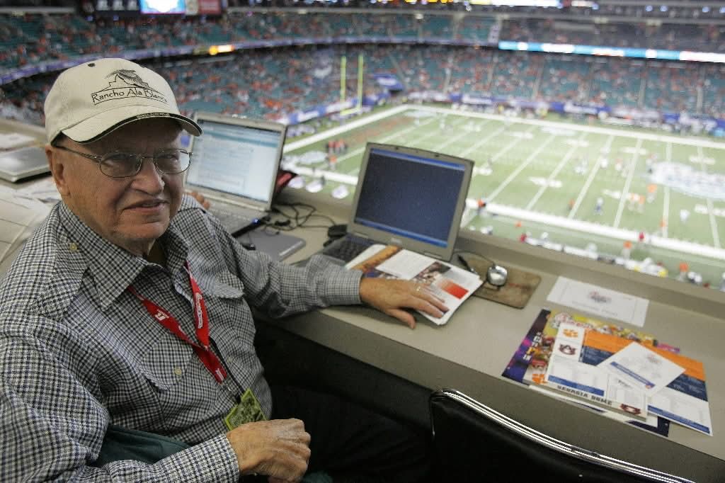 An older man sits at a computer in a press box at a football stadium, with the field and crowd visible in the background. 