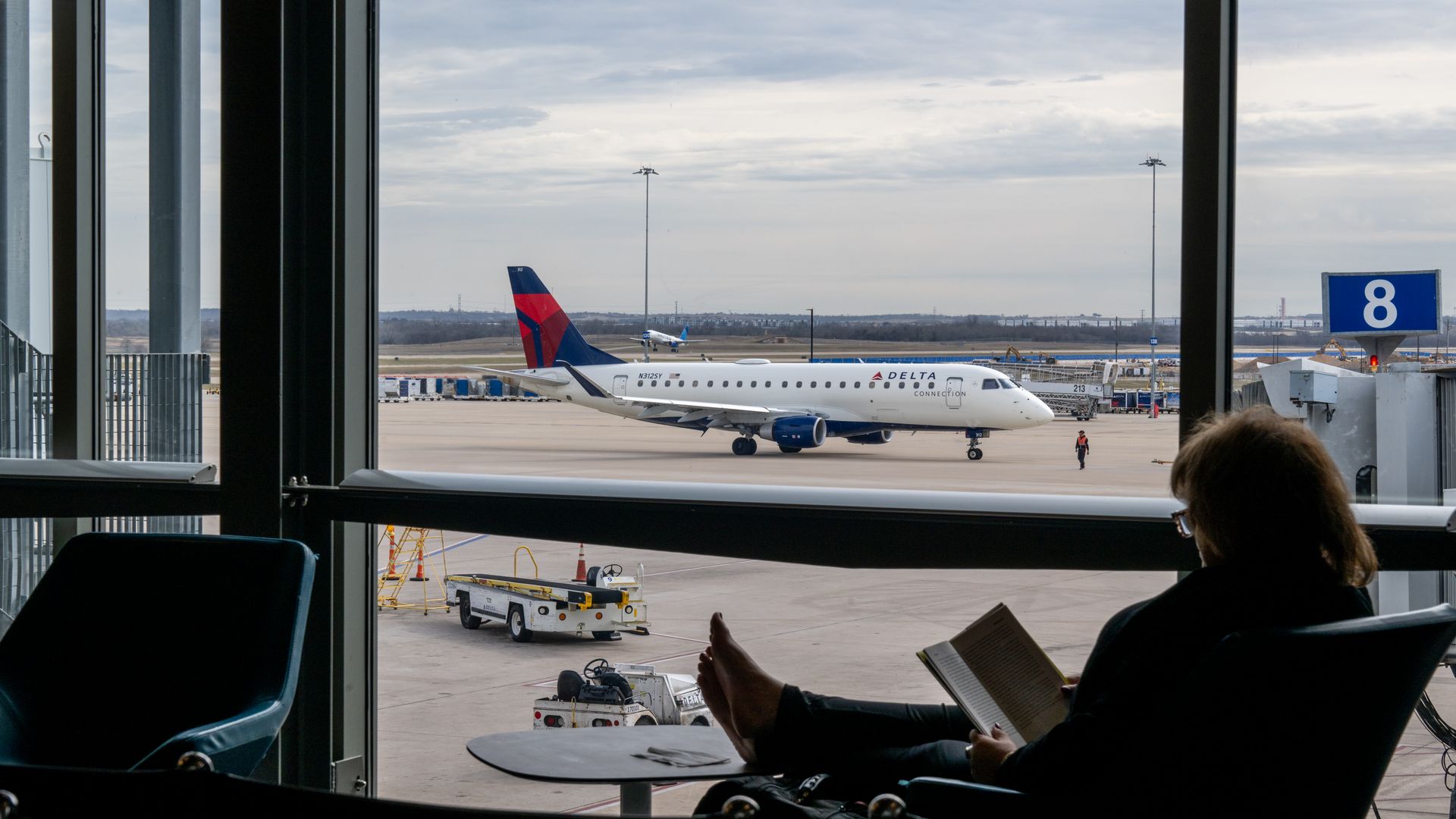 Person with glasses sitting barefoot in airport lounge reading, looking out large window at a Delta Connection airplane on the tarmac near gate 8 under a cloudy sky.