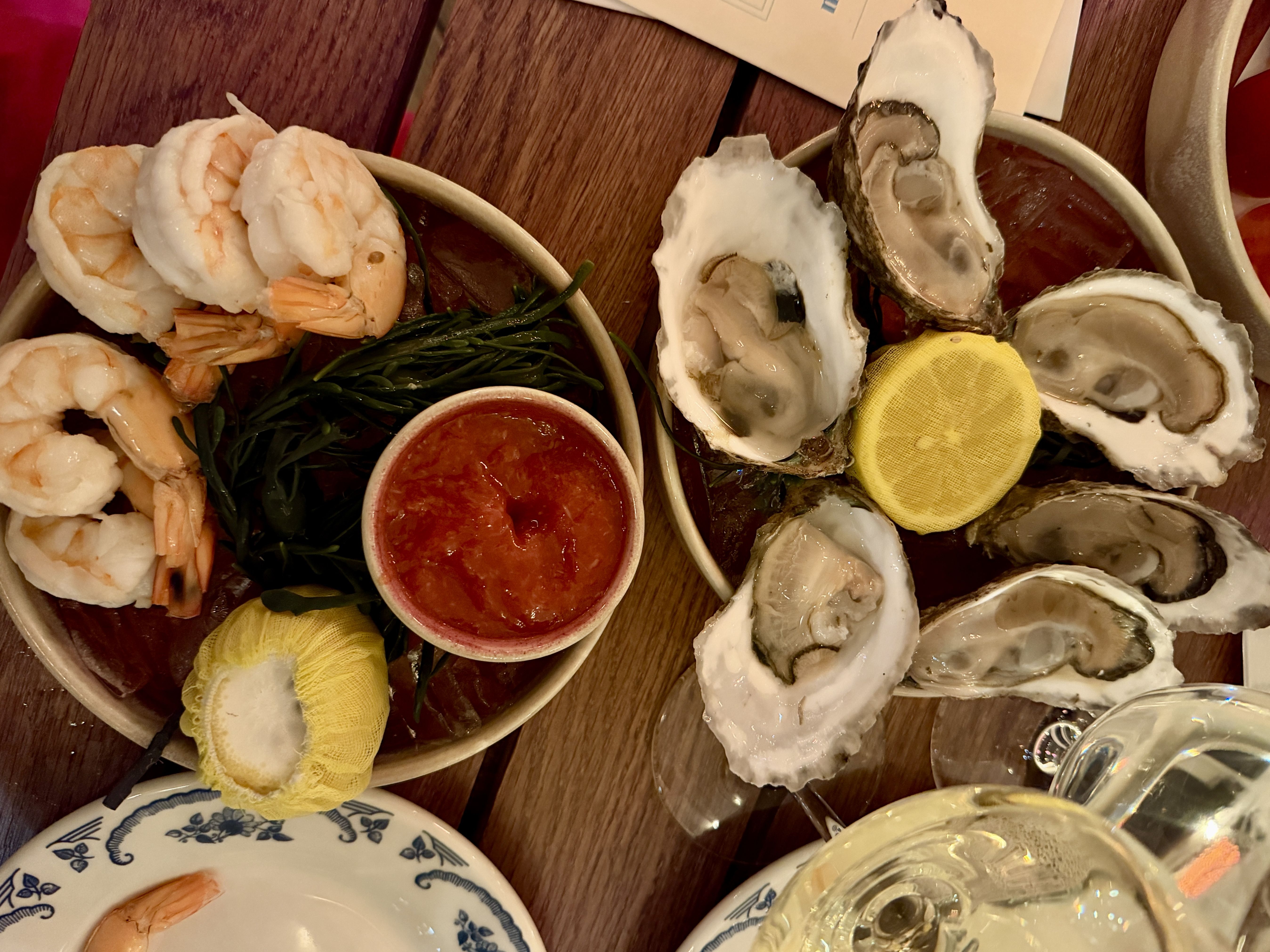 Plates with shrimp cocktail and raw oysters on the half shell, lemon halves covered with yellow mesh, cocktail sauce, seaweed garnish, and a glass of white wine on a wooden table.