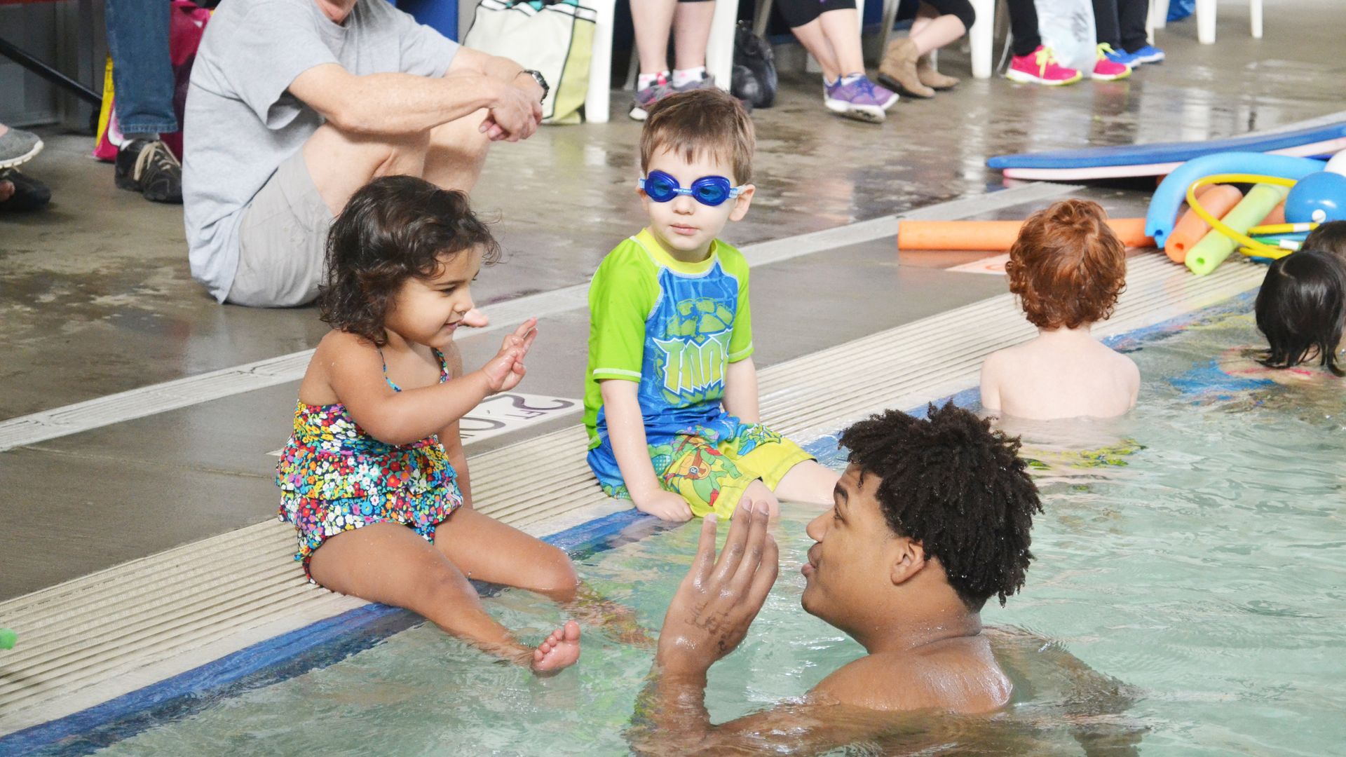 Two small children sit on the edge of a pool interacting with a swim instructor.
