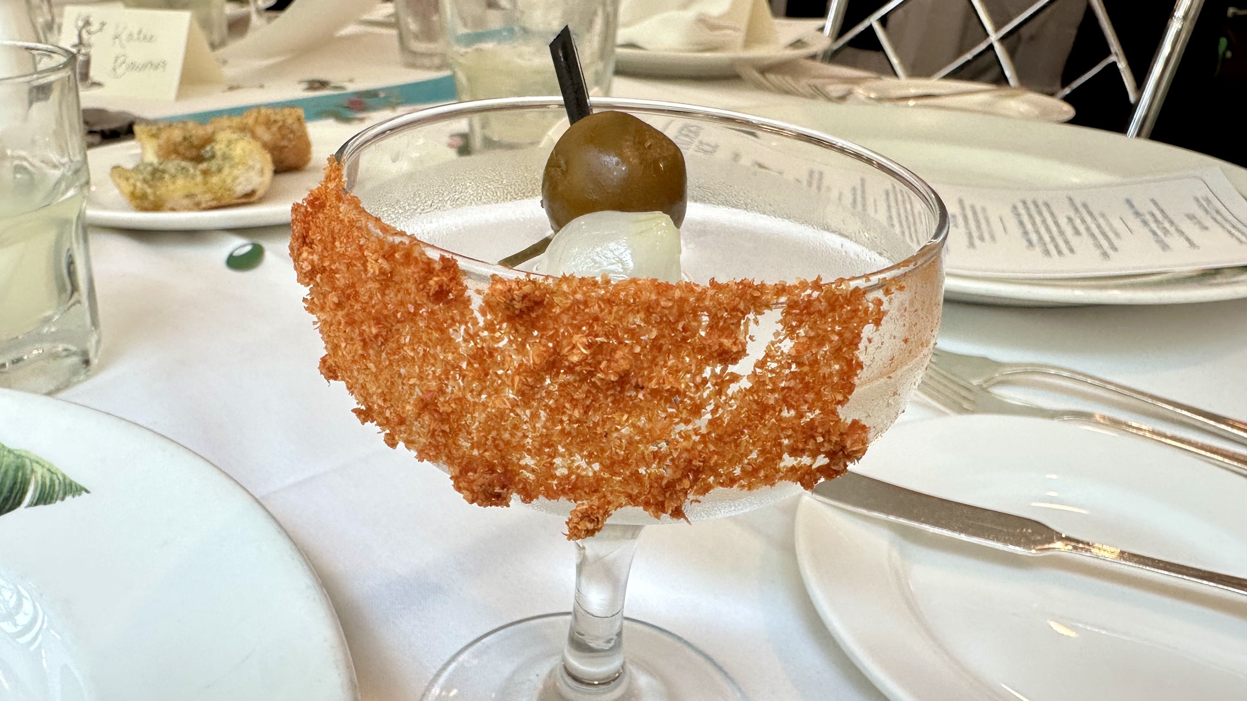 Cocktail glass with reddish spice rim, garnished with an olive and a pickled onion, on a white tablecloth with plates, a menu, and a small plate of bread in the background.