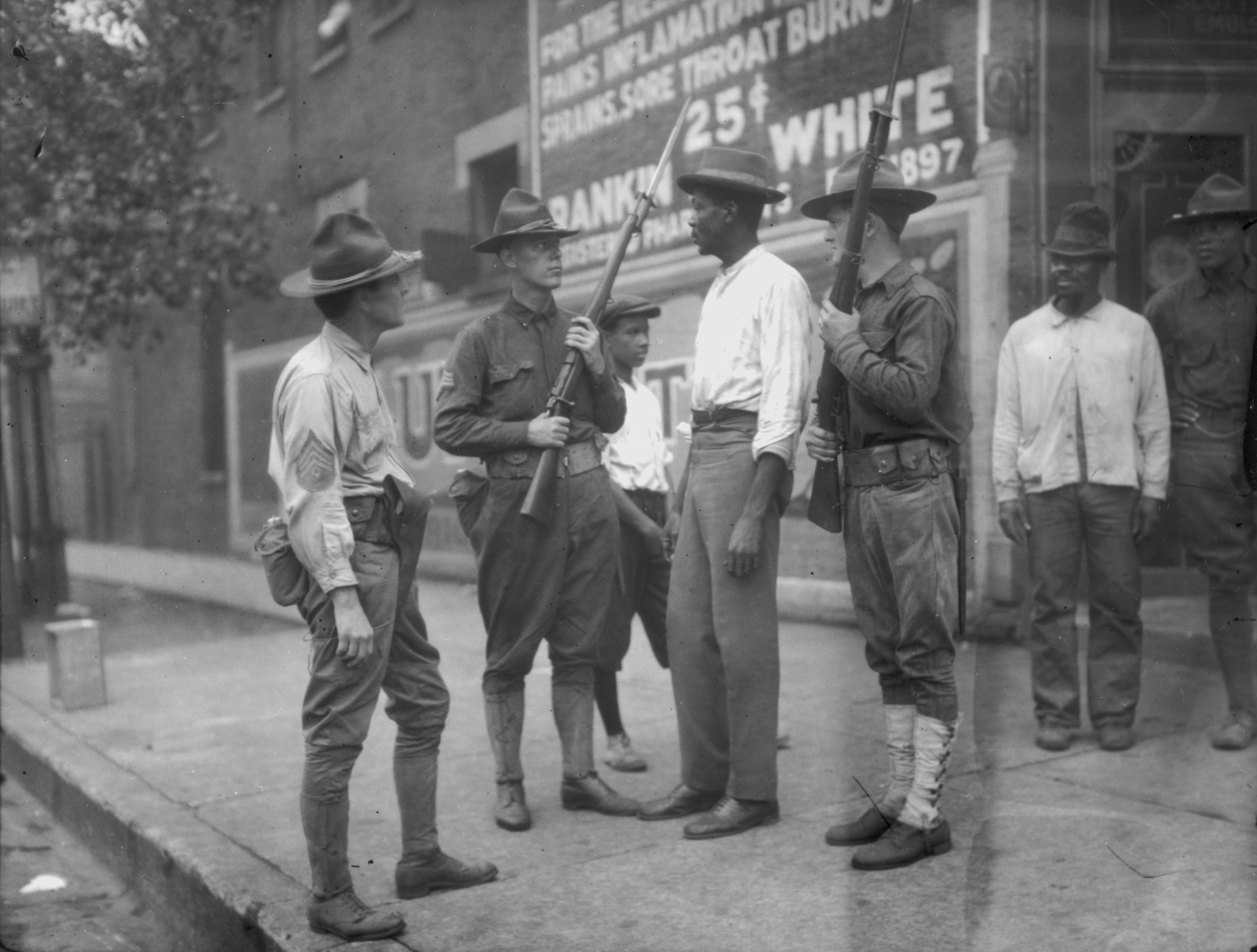 Black-and-white image of several soldiers in vintage uniforms with wide hats, standing on a sidewalk and talking, each holding rifles. A brick wall and a large sign are behind them.