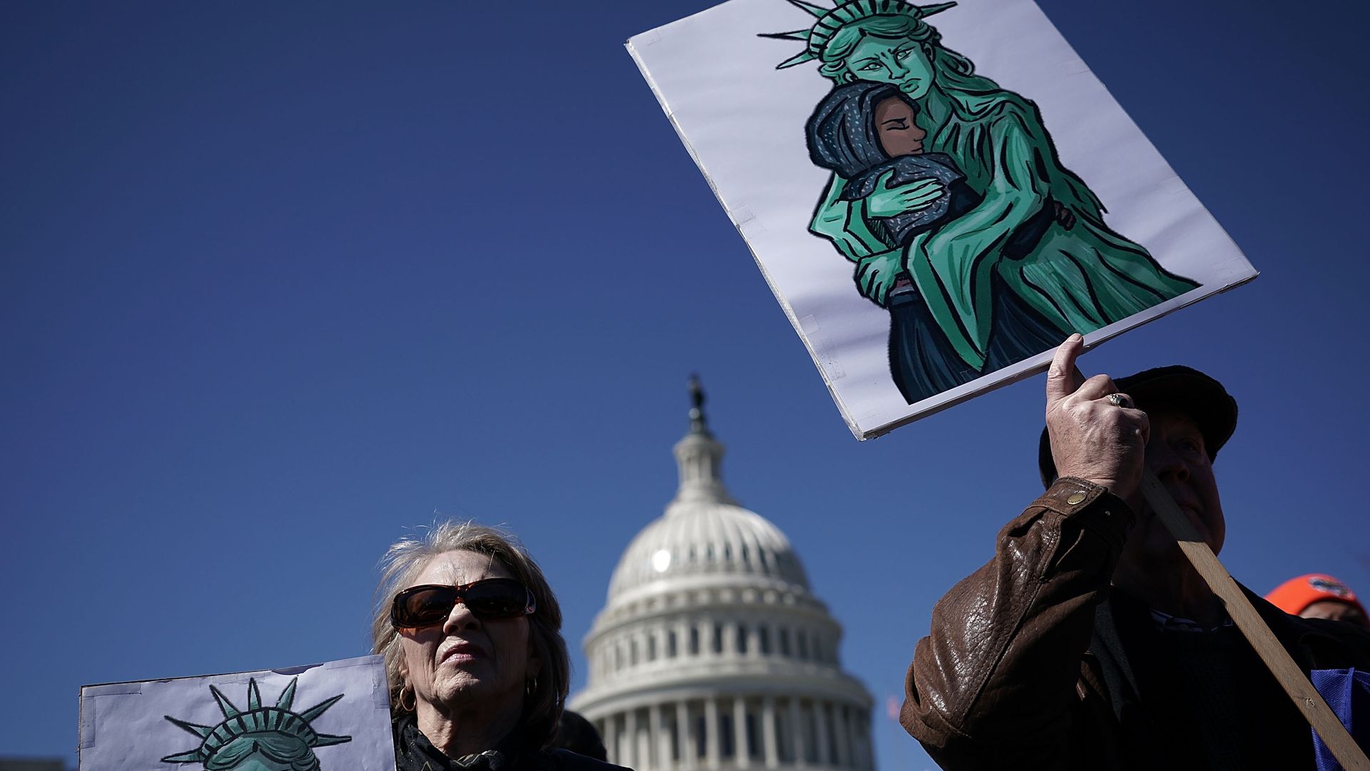 Two protestors holding signs of the statue of liberty hugging a woman with a hijab. The capitol building is in the background. 