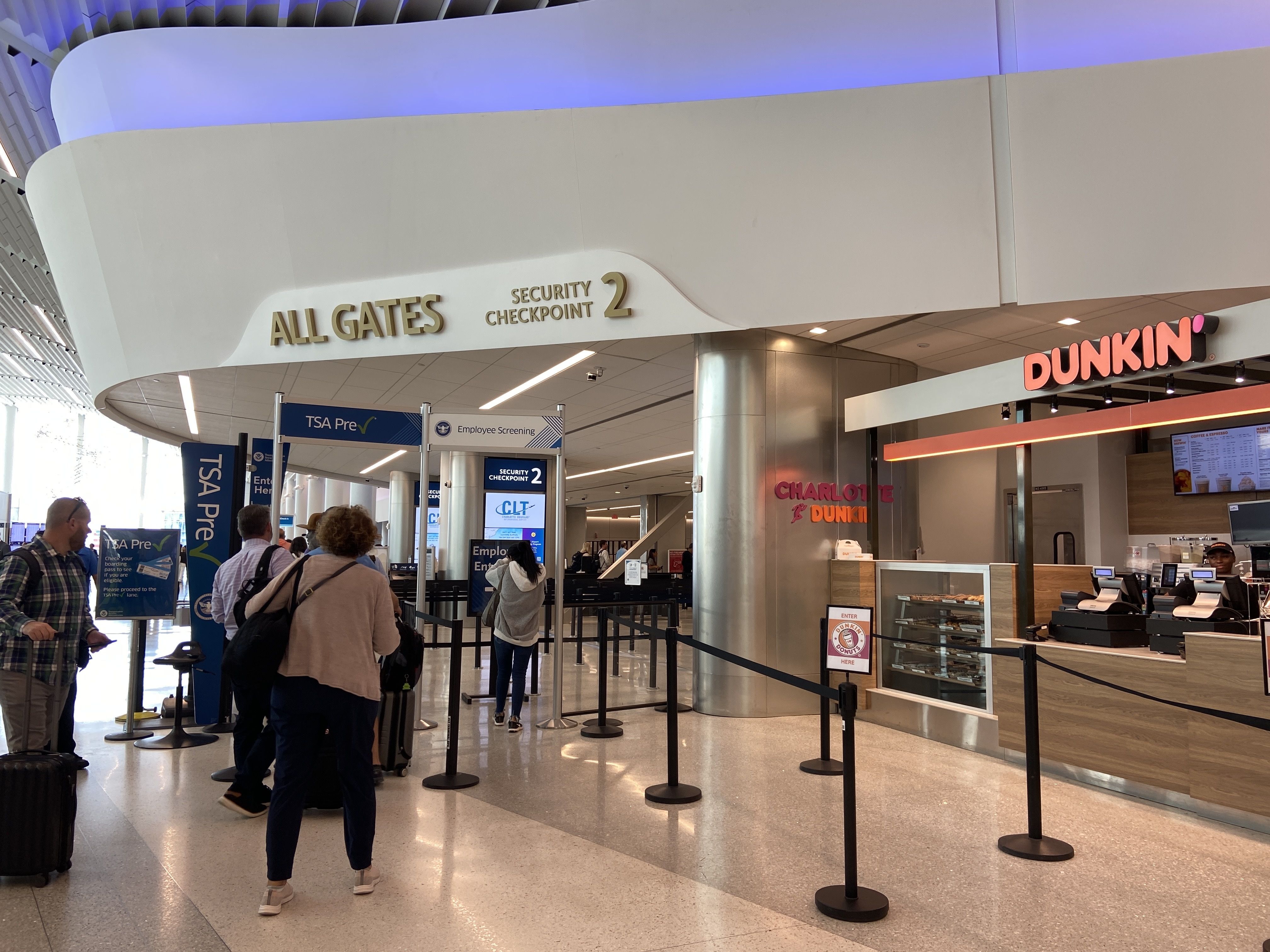 Travelers wait in line at airport Security Checkpoint 2 near TSA Pre area, with a Dunkin' coffee shop on the right and signage for all gates above; ceiling lit with blue lighting.