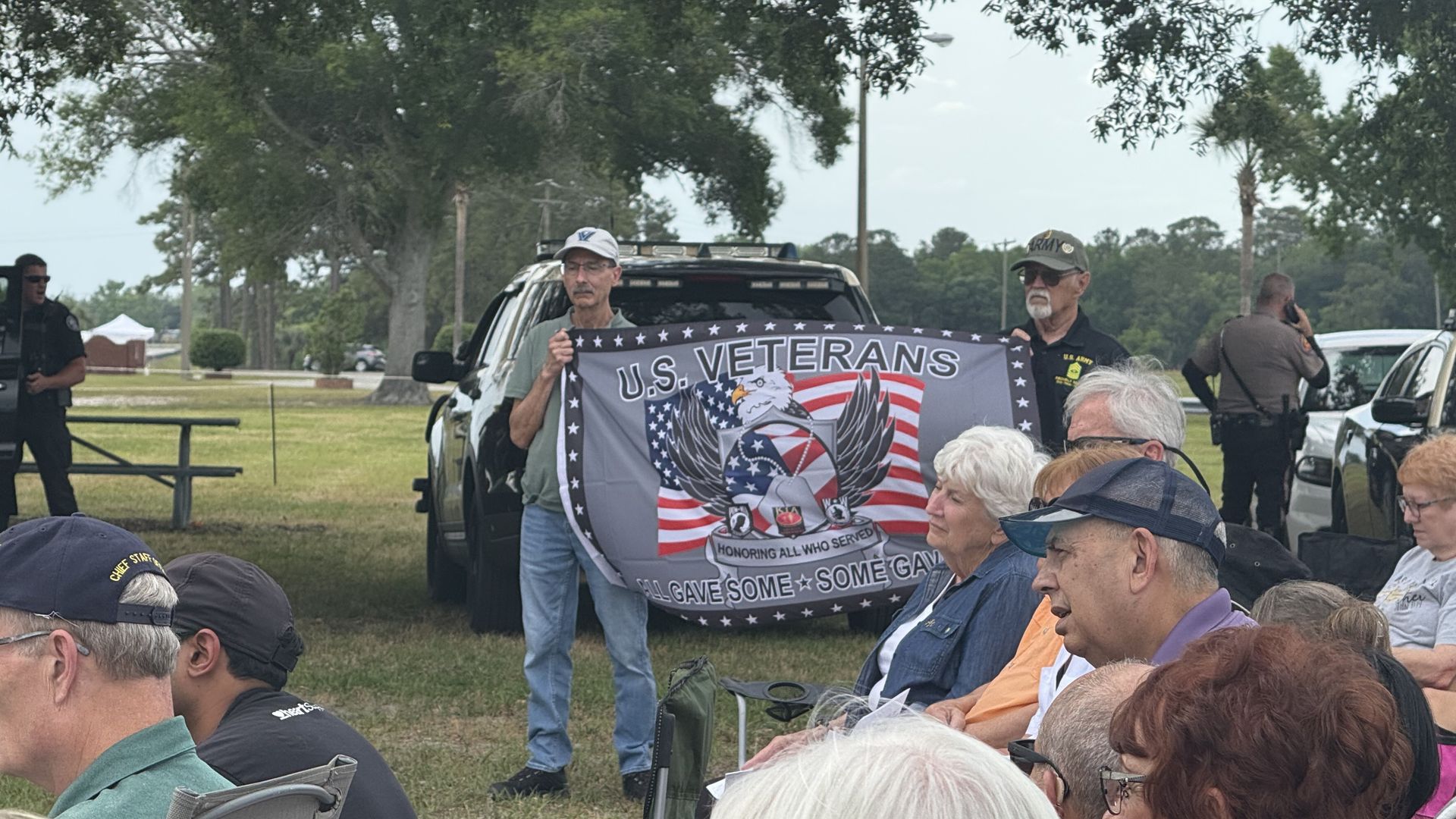 Two men stand in front of a black vehicle holding a U.S. Veterans flag with an eagle and American flag design. Several seated older adults watch attentively outdoors on a grassy area.