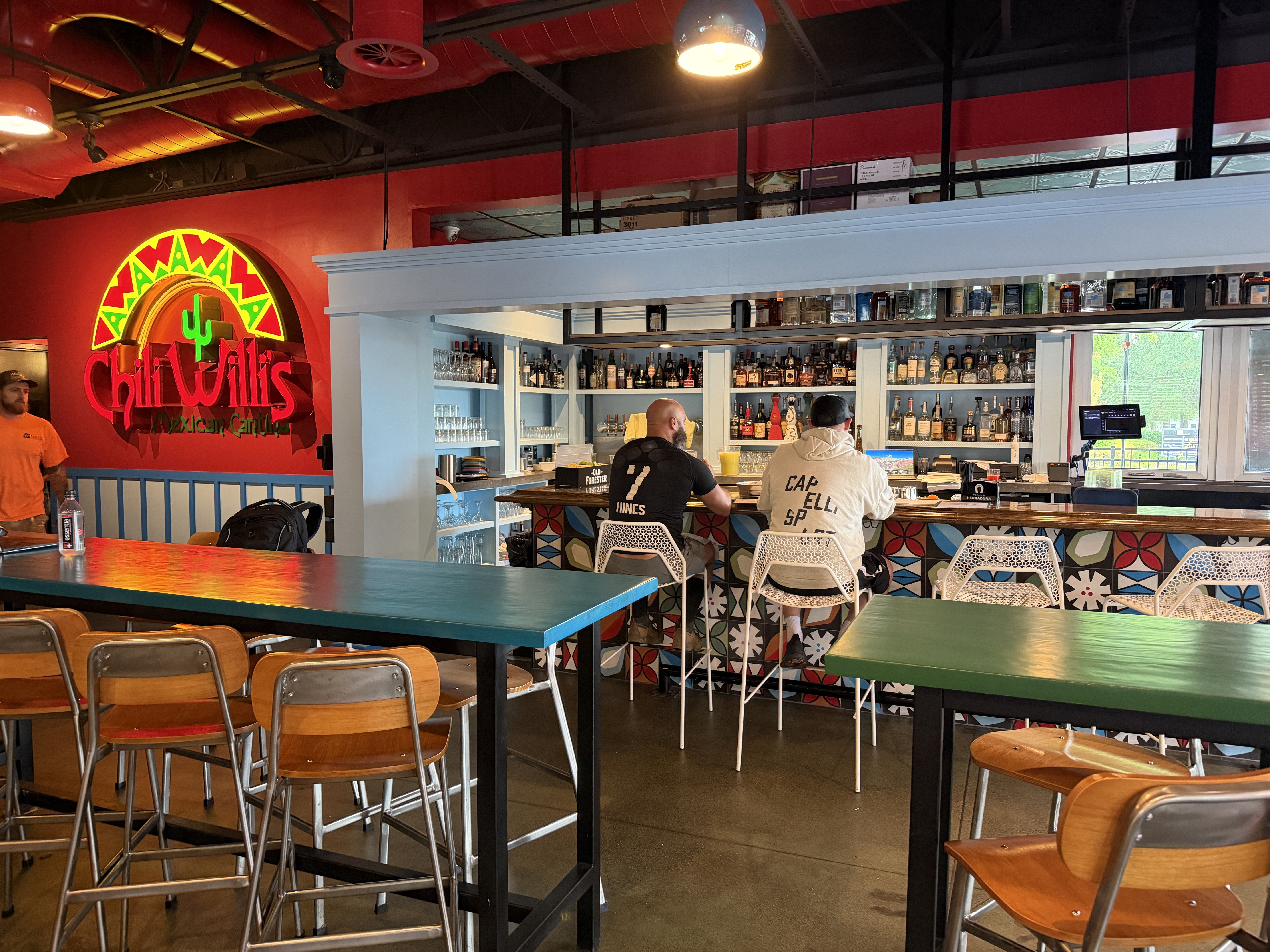 Colorful bar interior with a red wall neon sign reading "Chili Will's Mexican Cantina", blue liquor shelves, turquoise tables, and customers seated at the bar.