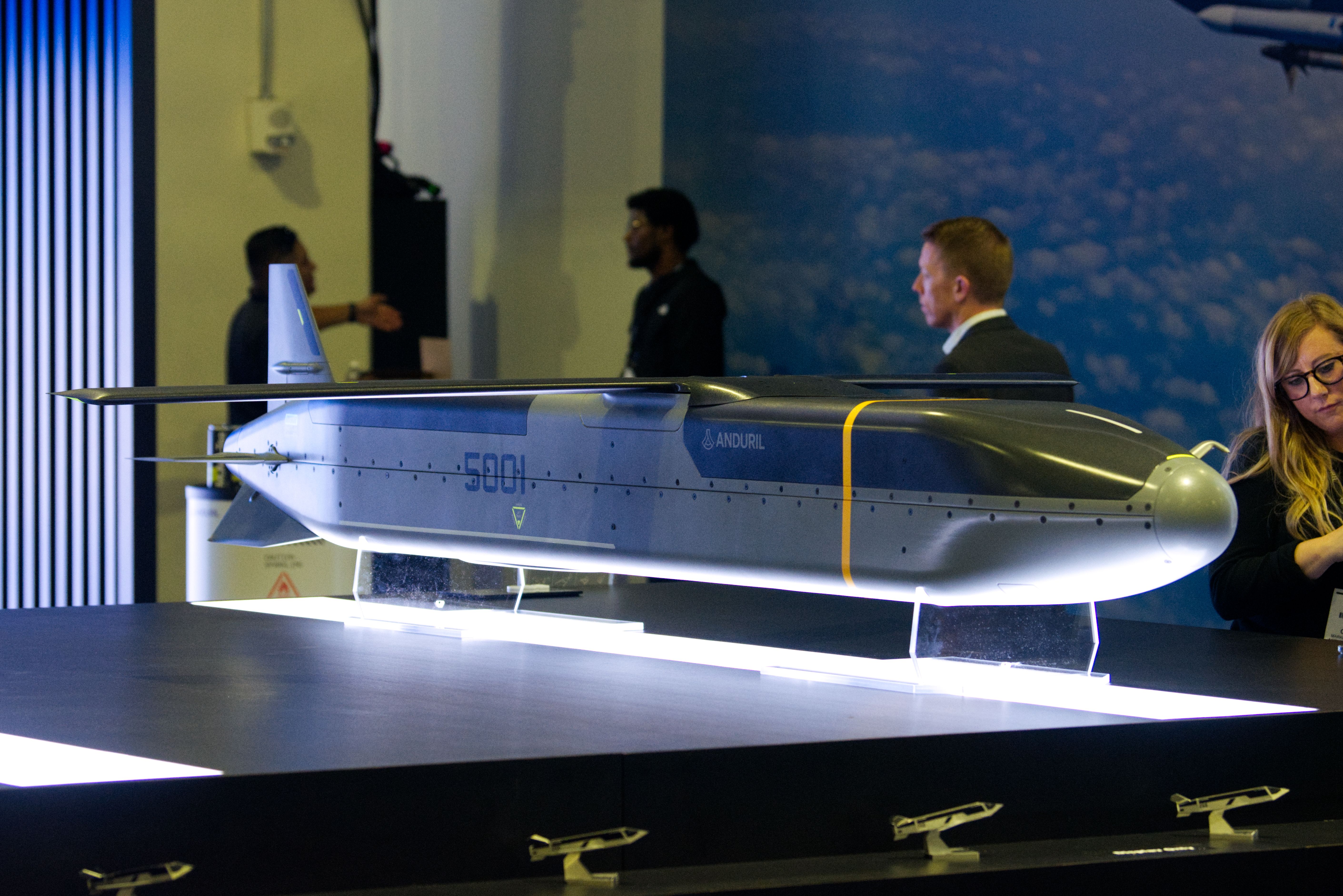 A black-and-grey missile sits at a defense tradeshow booth. It is lit from the bottom. People pass by.