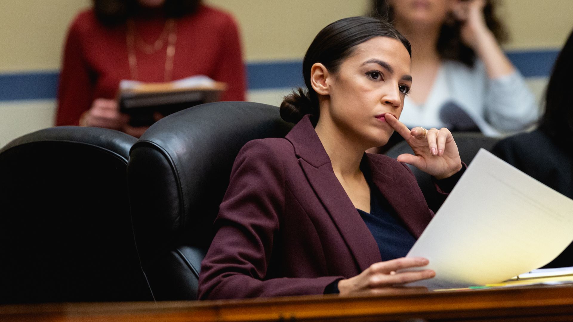 In this image, Alexandria Ocasio-Cortez sits and listens to Cohen's testimony while holding papers.