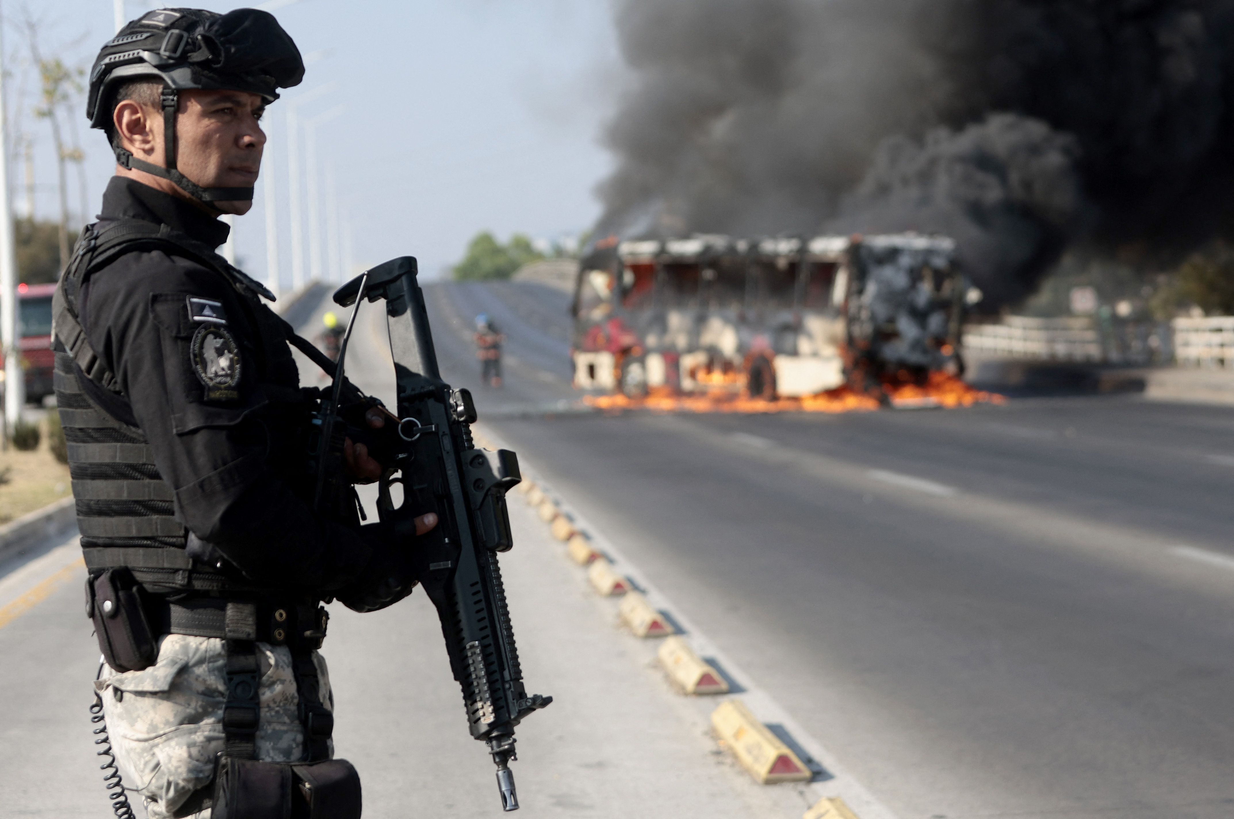A law enforcement officer stands near a burning bus in Zapopan, Mexico, after it was set on fire by a cartel yesterday.