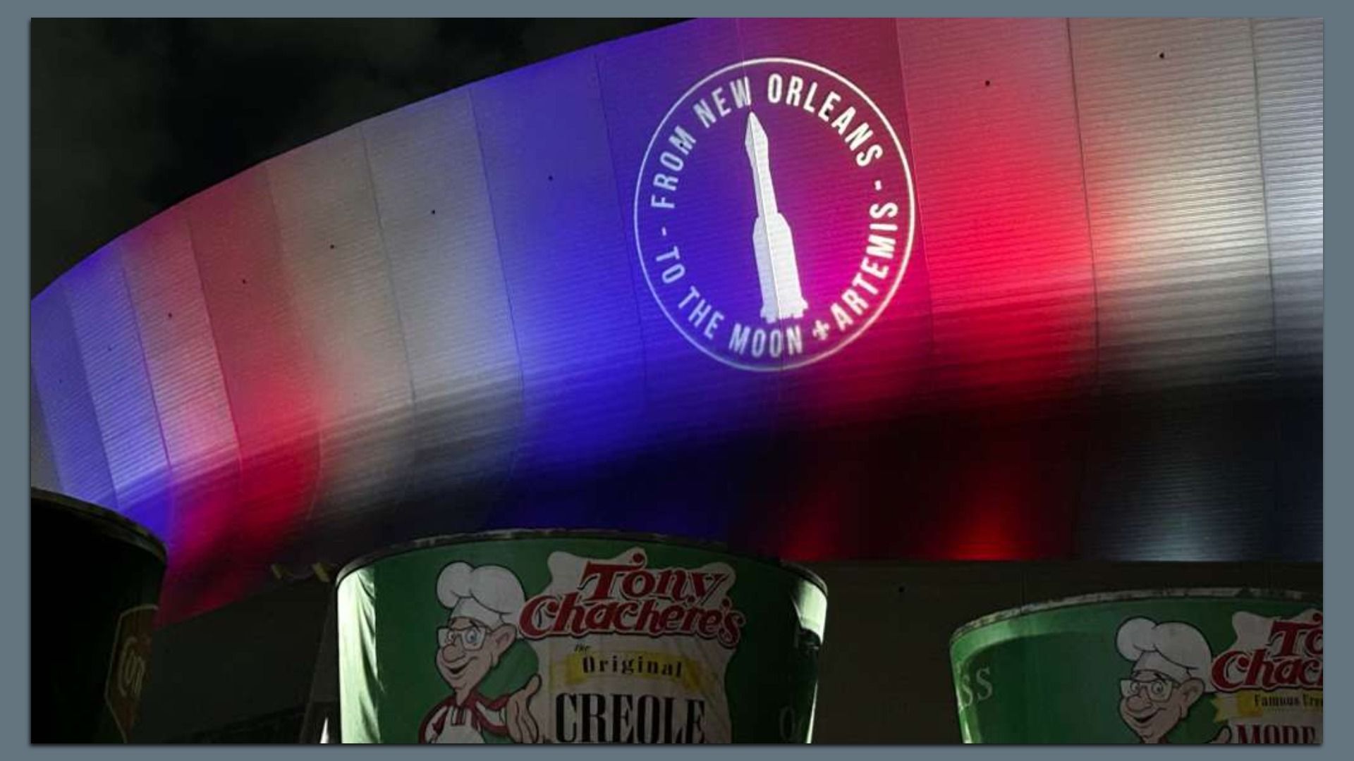 The Caesars Superdome lit in blue, white and red gradients features a circular logo with a white rocket and the text "From New Orleans to the Moon & Artemis". In front are green Tony Chachere's Creole seasoning containers.