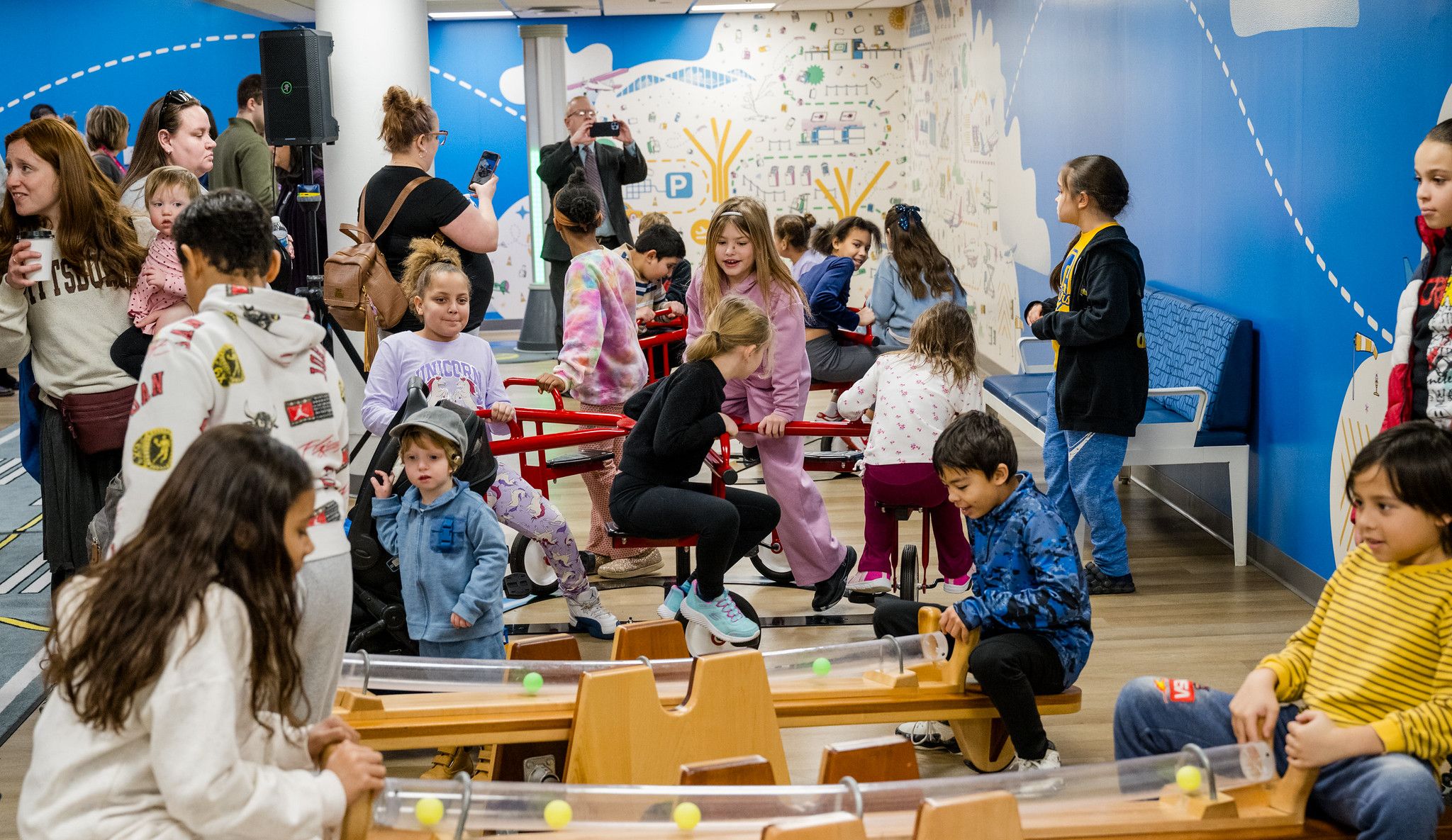 Group of children and adults in a colorful playroom with blue walls, wooden floor, and interactive play equipment. Kids engage with toys and each other, while some adults observe and take photos.