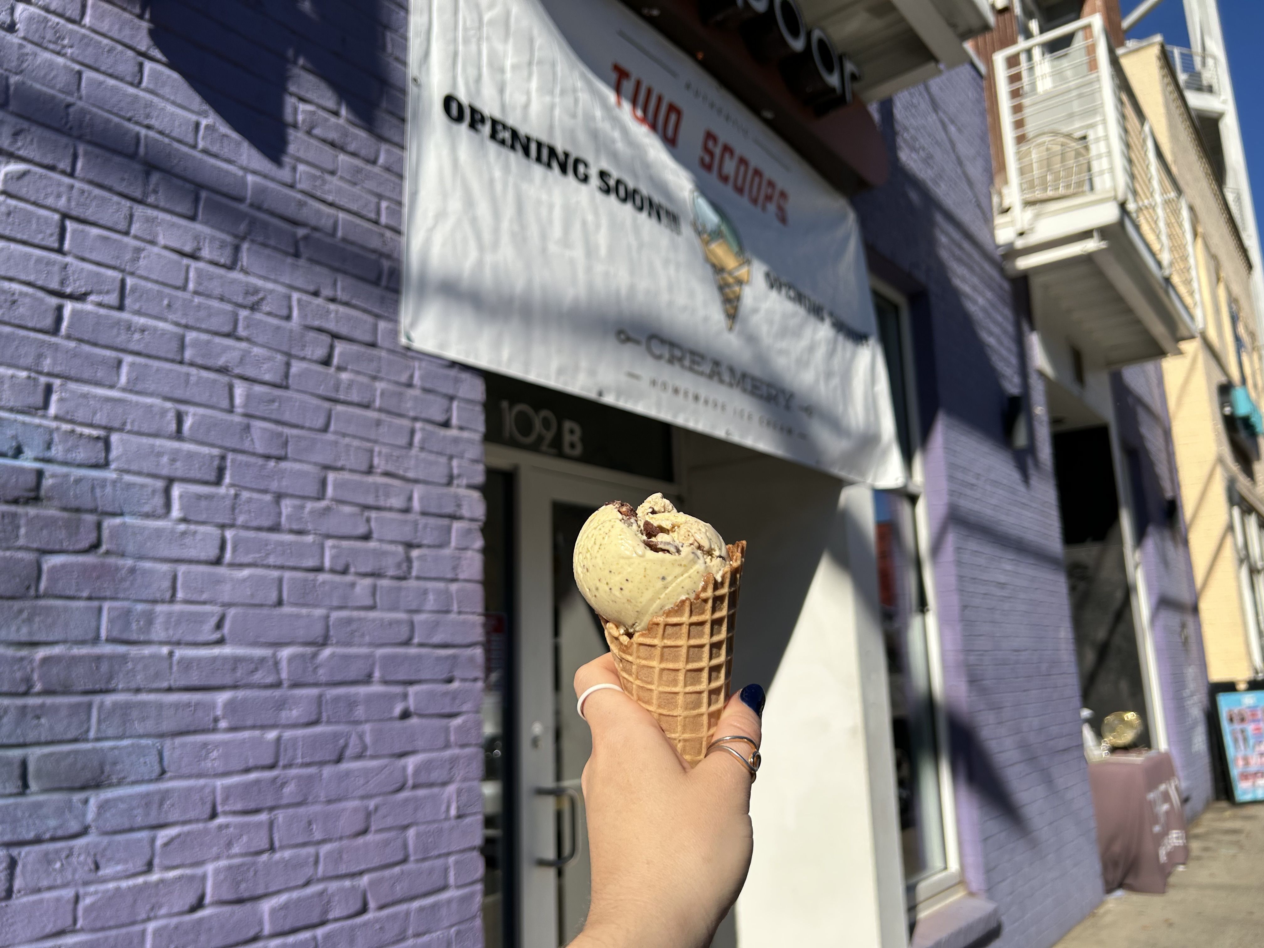 Hand holding a waffle cone with two scoops of light brown ice cream in front of a purple brick building. A sign above reads "Two Scoops Opening Soon".