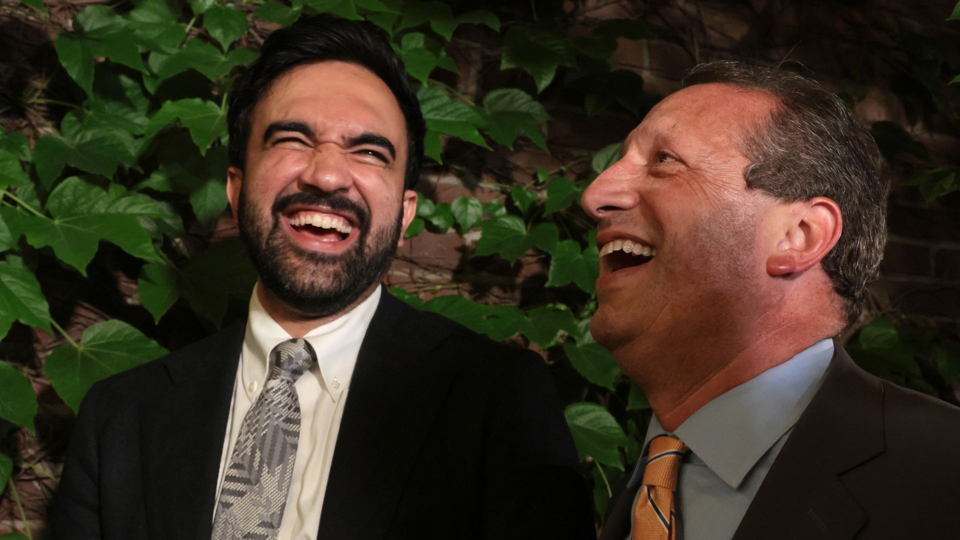Two men in suits laughing outdoors, left man with dark hair and beard, right man with gray hair, ivy-covered wall in the background.