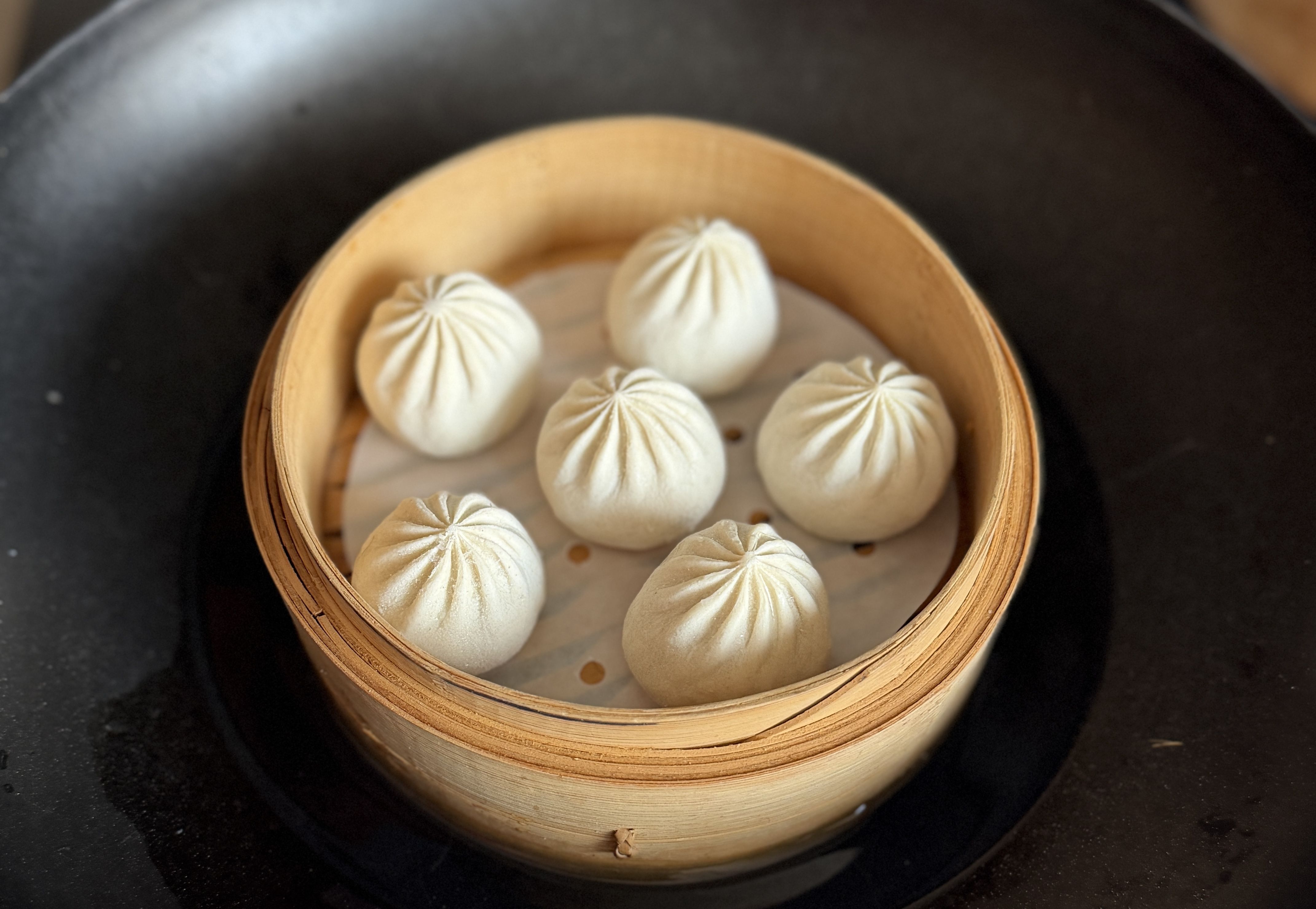 Six white steamed dumplings with pleated tops in a round bamboo steamer basket on a black surface.