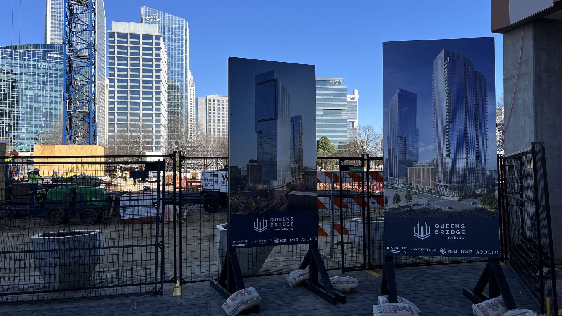 Construction site in a city with a tall crane and workers, two large signs display modern skyscraper renderings labeled "Queens Bridge Collective" against a clear blue sky.