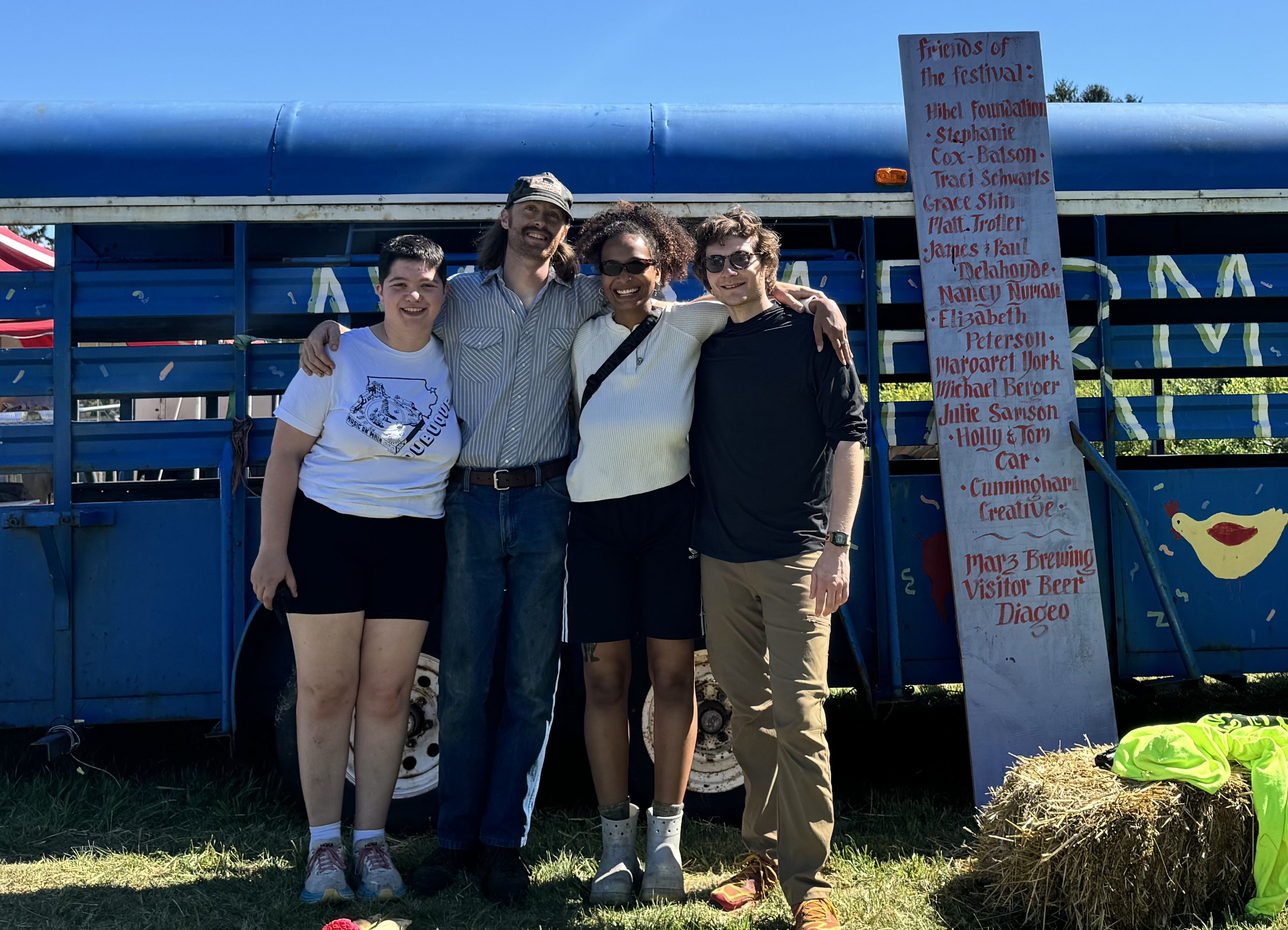 Four friends pose with arms around each other in front of a bright blue carnival ride; a tall sign listing red names stands to the right under a clear sky.