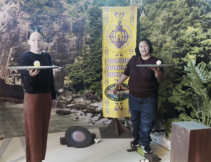 Two people stand on beams holding up a tray balancing a while ball on it as part of the "ball roller" challenge at the "Survivor"-themed cafe in downtown Boston.