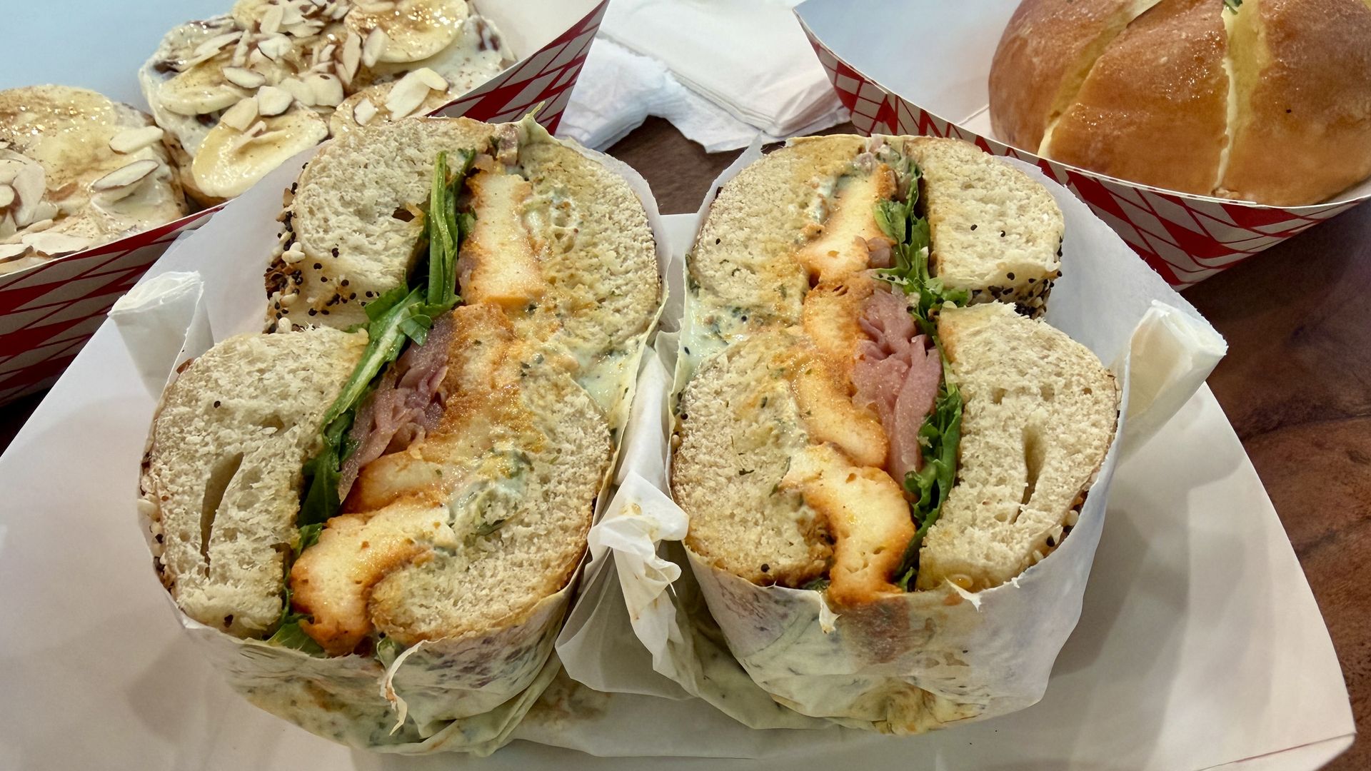 Two halves of a seeded sandwich with greens and a creamy herb sauce, wrapped in parchment, sit on a white tray; in the background are almond-topped pastries and a round loaf.