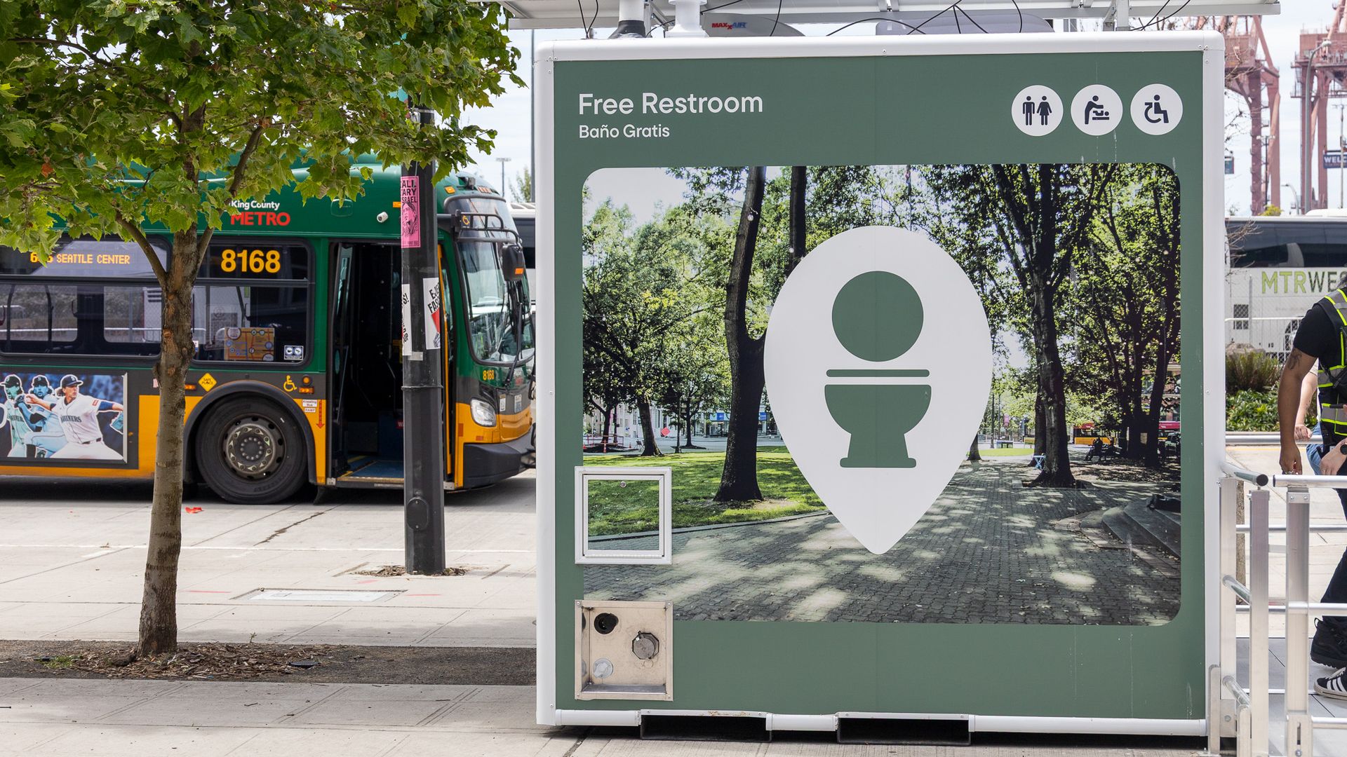Green sign reads "Free Restroom" with "Baño Gratis" and icons for men and wheelchair. Behind it, a green-yellow Metro bus passes by; trees and cranes are visible in the background.