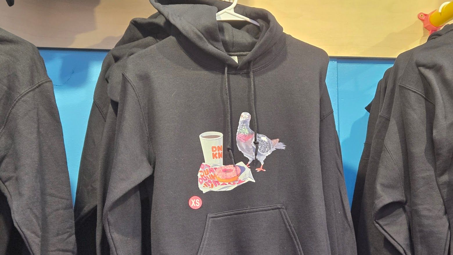 Black hoodie with a graphic of a pigeon standing next to a coffee cup and pink donut on a wrapper, hanging among similar hoodies against a blue and beige background.