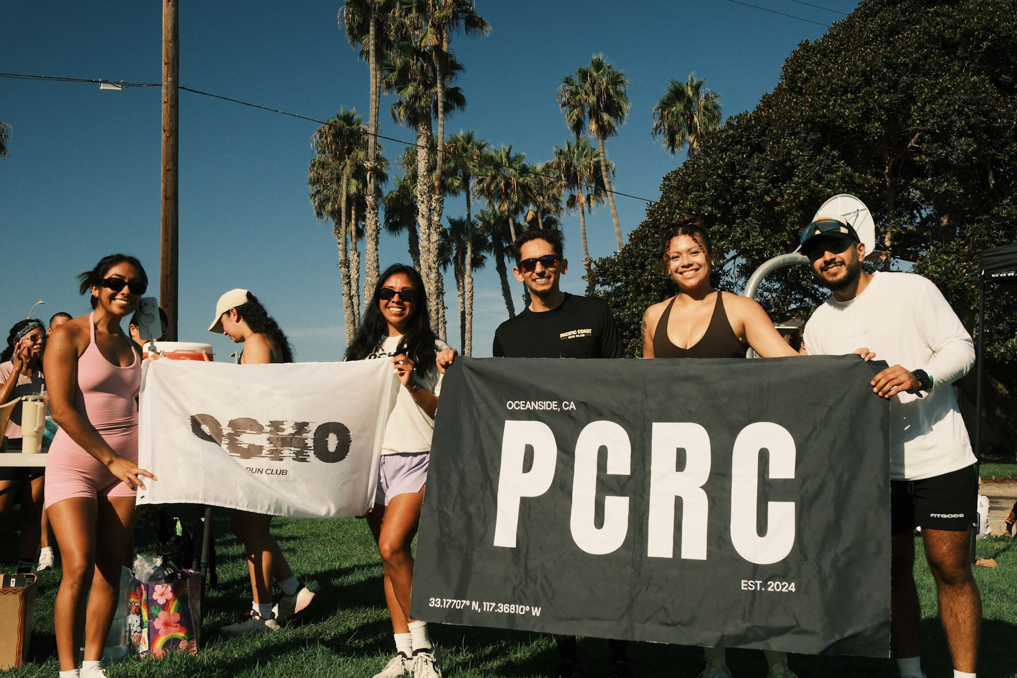 Group of friends posing at an outdoor event, holding two banners. Sunny day with tall palm trees and a deep blue sky behind them; participants wear sunglasses and casual summer outfits.