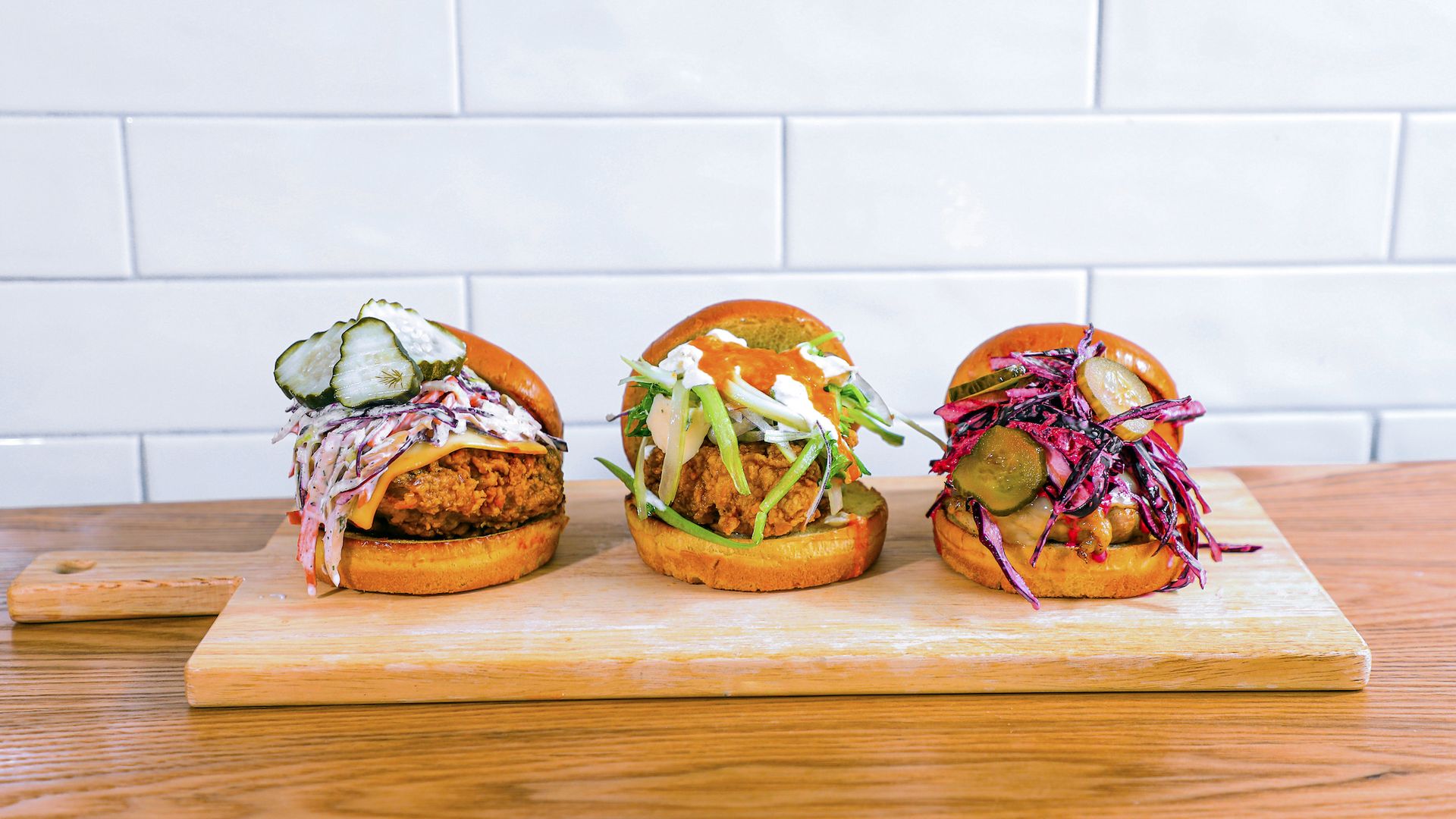 Three different gourmet fried chicken sandwiches topped with pickles, coleslaw, and sauces, served on a wooden board against a white subway tile background.