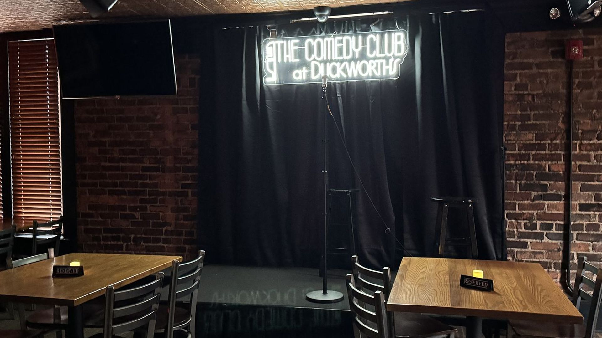 Dim comedy club stage with a neon sign above a black curtain; microphone on a stand center stage. Brick walls flank the stage, with wooden tables and chairs in foreground and window blinds on left.