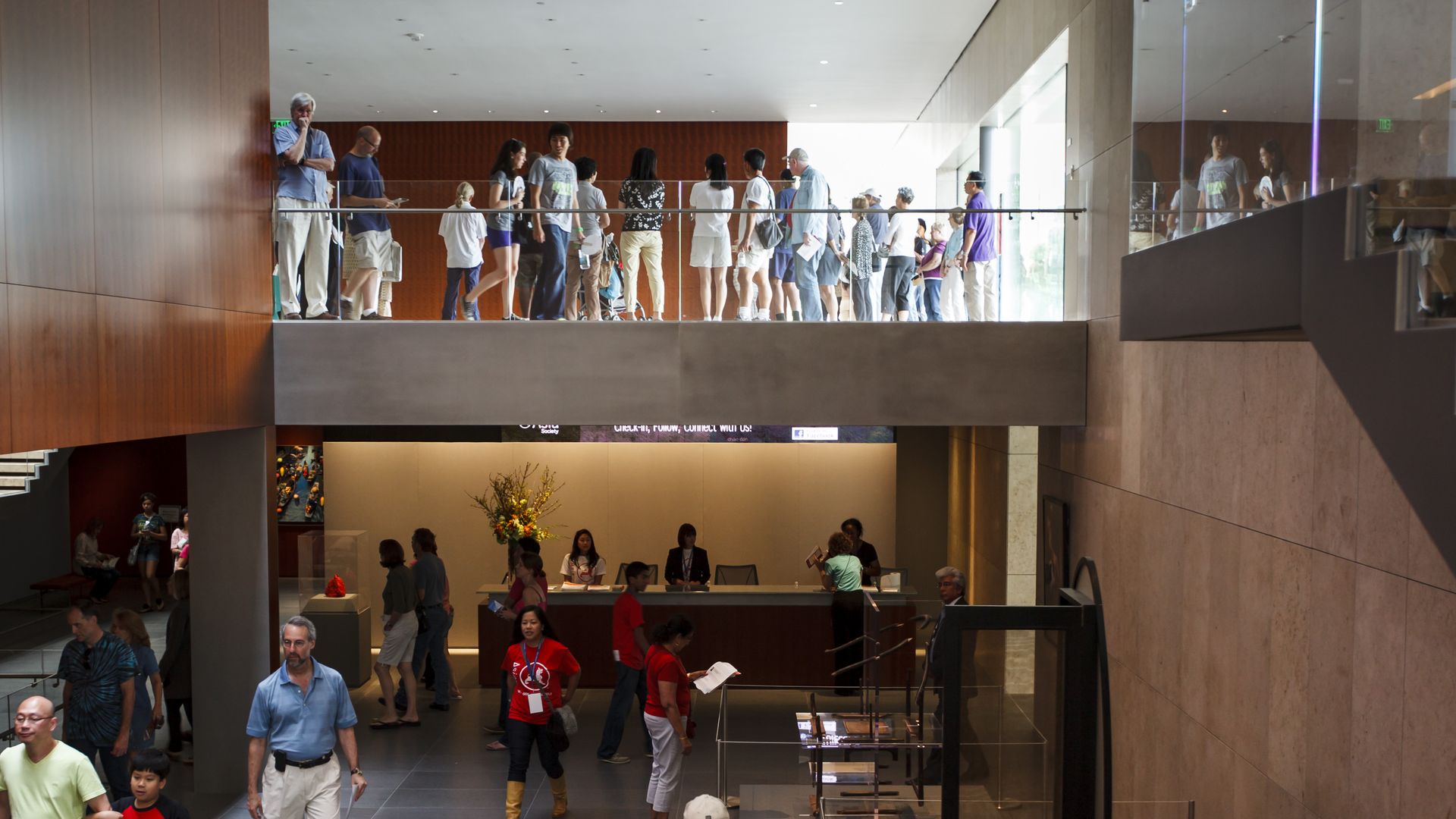 Wide interior atrium with a crowd lined along a glass railing on the mezzanine. Wood-paneled wall left, marble wall right, and a reception desk below as visitors walk and chat.