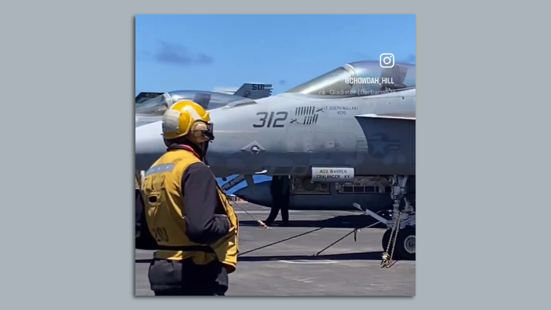 A person clad in yellow protective gear looks at grey fighter jets aboard an aircraft carrier. The frame is tight, but blue sky can be seen above.
