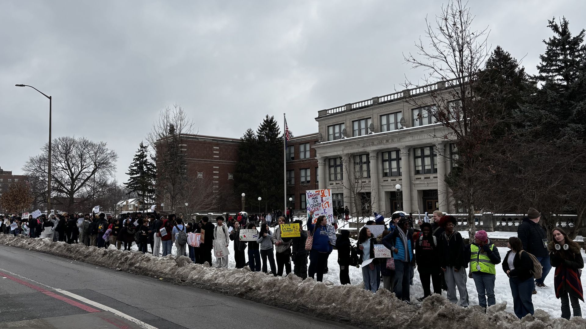Large group of people standing in a snowy street holding protest signs outside a neoclassical building under gray cloudy sky.