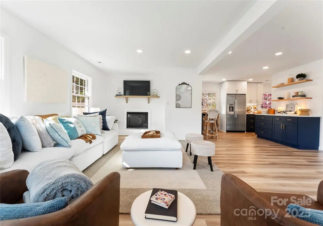 Bright open living room with white walls, white sectional sofa with blue cushions, brown armchairs, wooden floor, navy blue kitchen cabinets, and stainless steel refrigerator.