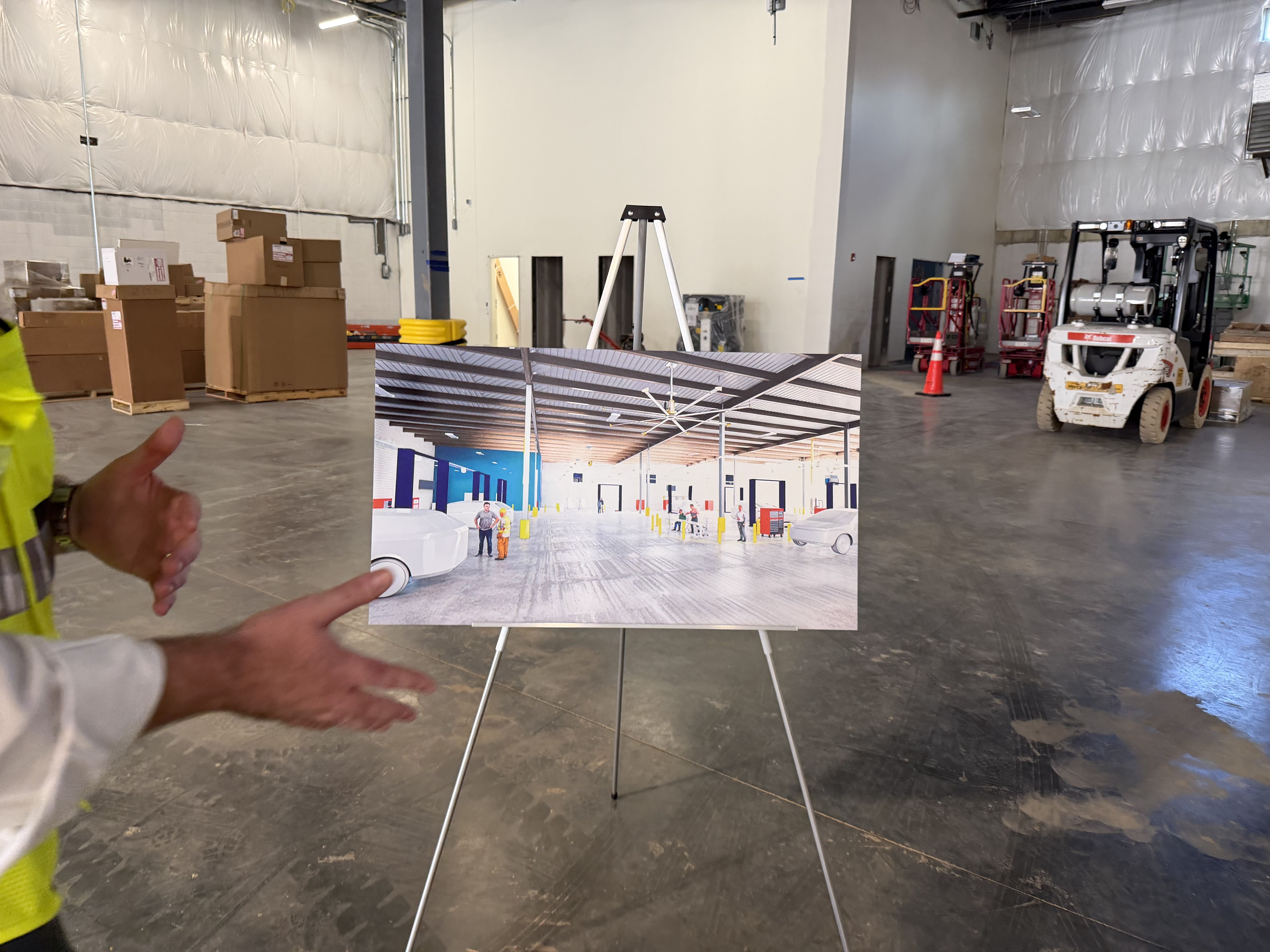 Industrial warehouse with white walls, stacked boxes on the left, a forklift and orange cones on the right. A person in a yellow safety vest points to a photo board showing a finished interior space.