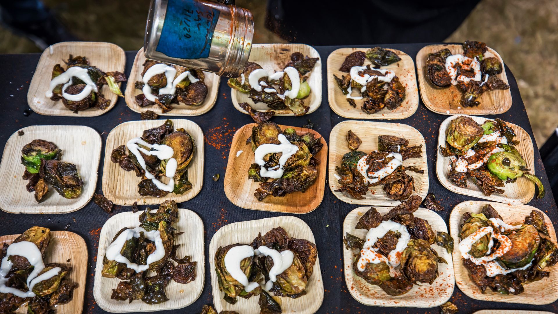 A row of small wooden plates on a dark table, each with roasted greens and meat topped with white sauce; a metal jar tilts to pour sauce over several plates.