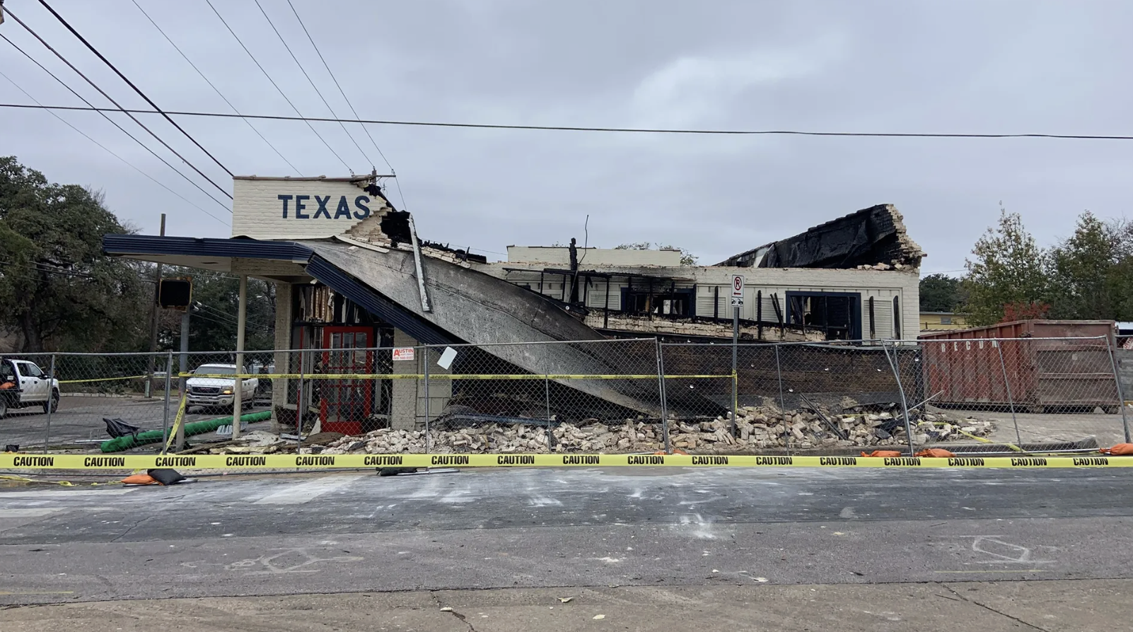Damaged building with collapsed roof and burned walls, marked "TEXAS", surrounded by caution tape and fencing, under a gray sky with some trees and vehicles nearby.