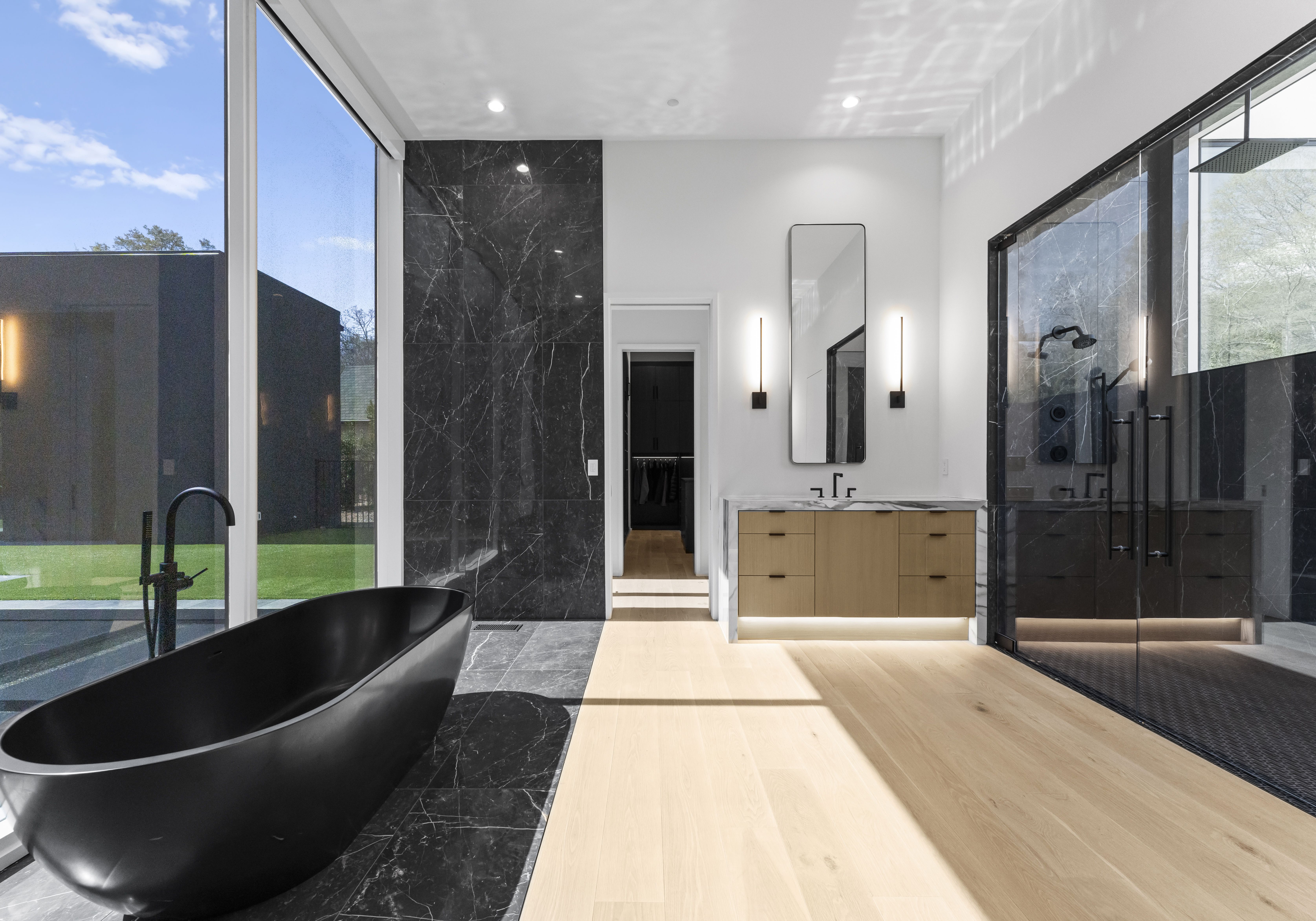 Modern bathroom with a matte black freestanding tub, black marble tiles, light wood floor, double glass shower, and wooden vanity with tall vertical mirror and two vertical lights.