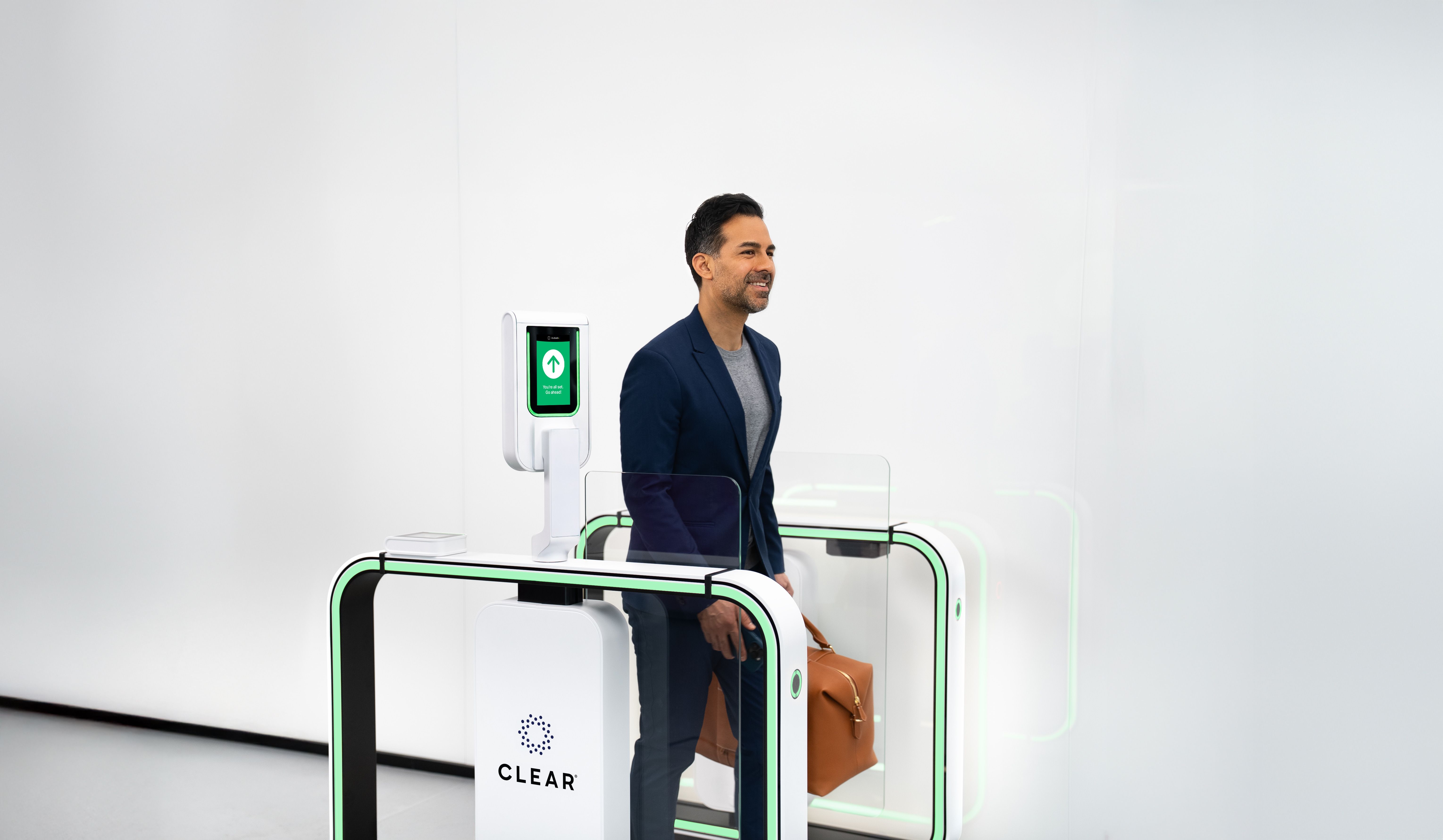 Man in a navy blazer walking through a CLEAR security gate with a green arrow on the screen, carrying a brown leather bag, in a sleek, white modern space.