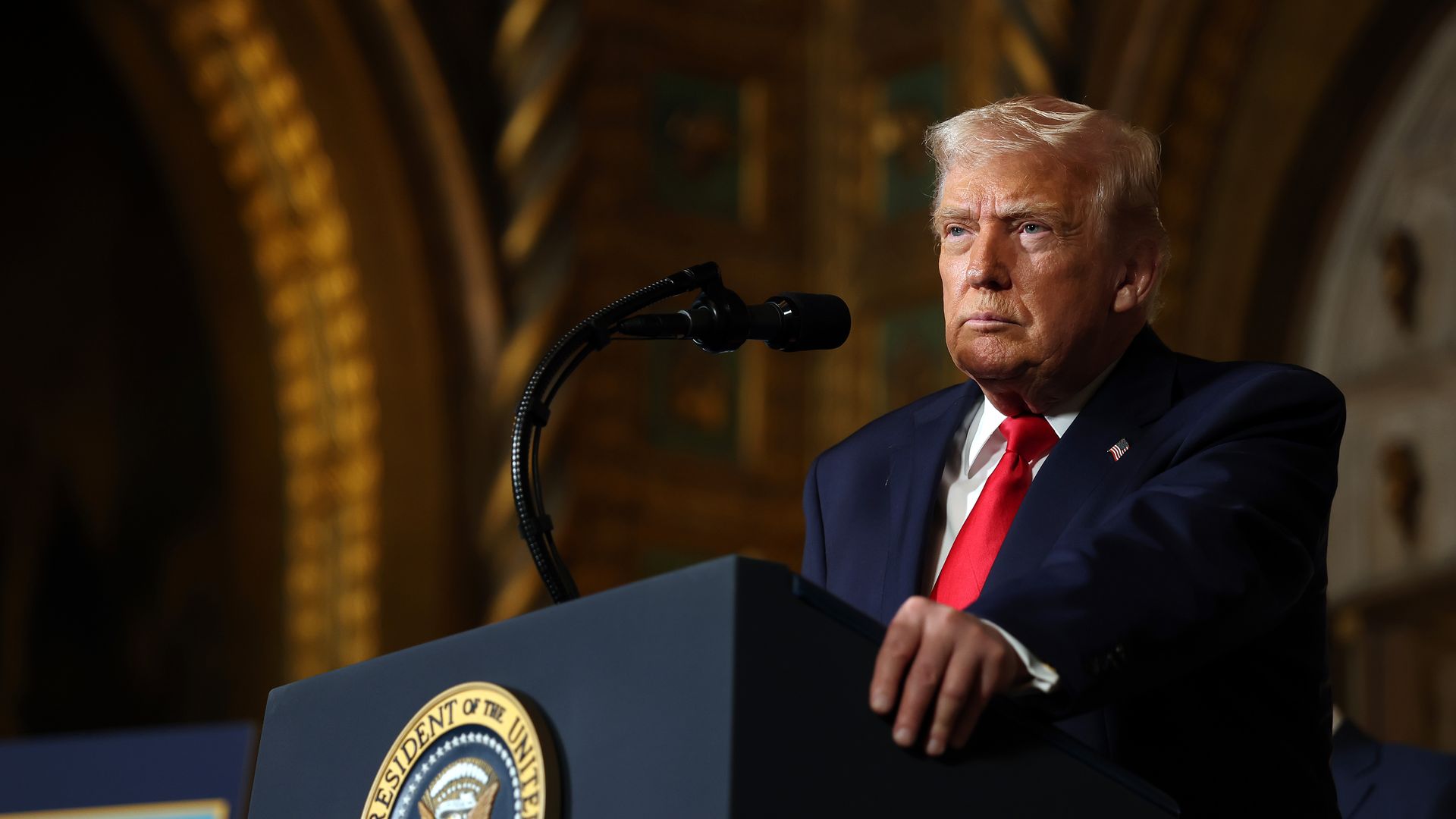 Trump with a straight face while standing at a podium wearing a navy suit jacket with an American flag pin, white collared shirt and red tie