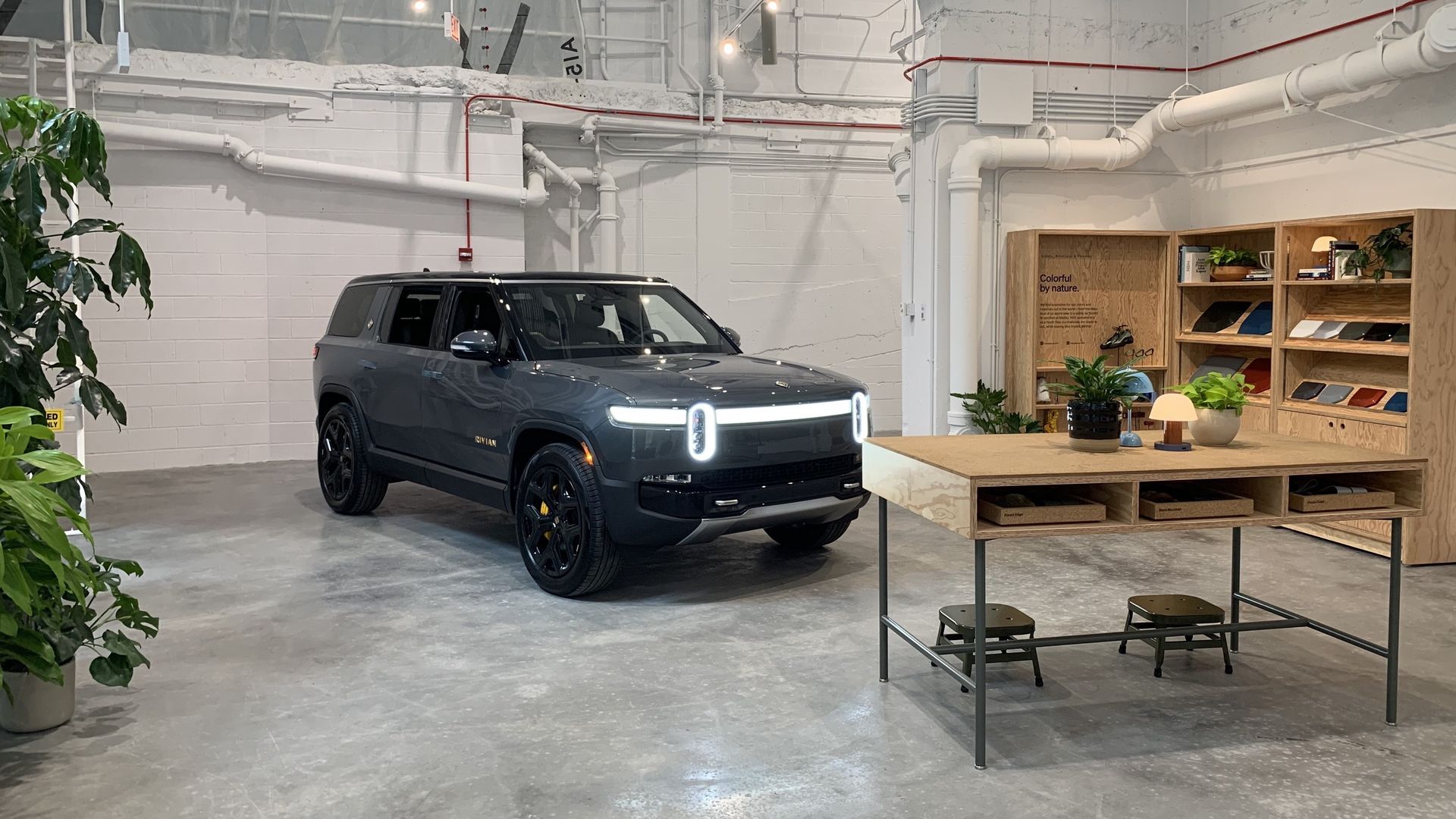 Blue truck next to greenery and behind a wood desk, next to a bookshelf in a concrete warehouse.