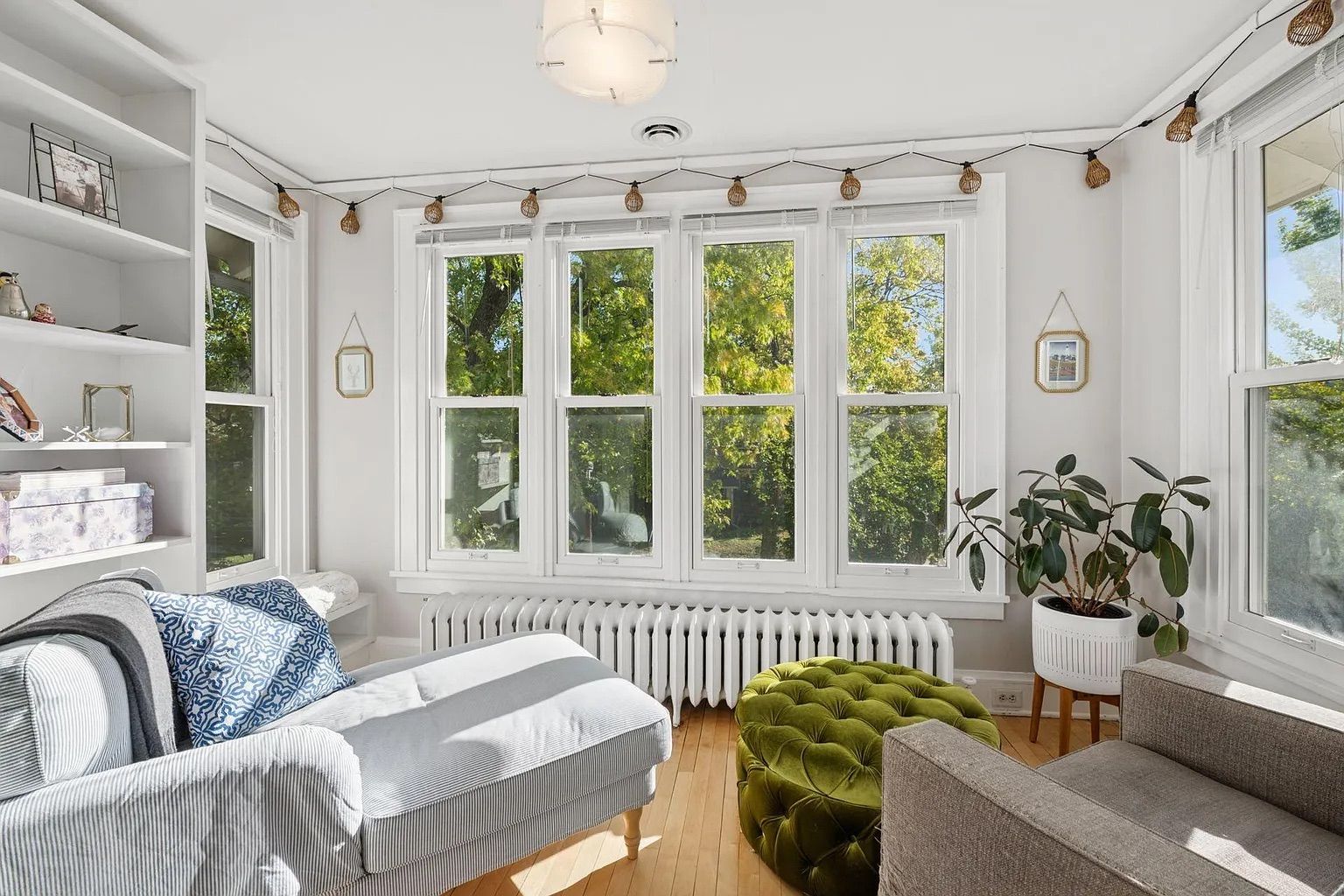 Bright living room with five large windows, a light gray chaise lounge with blue patterned pillow, green tufted ottoman, gray armchair, potted plant, white walls, and wood floor.