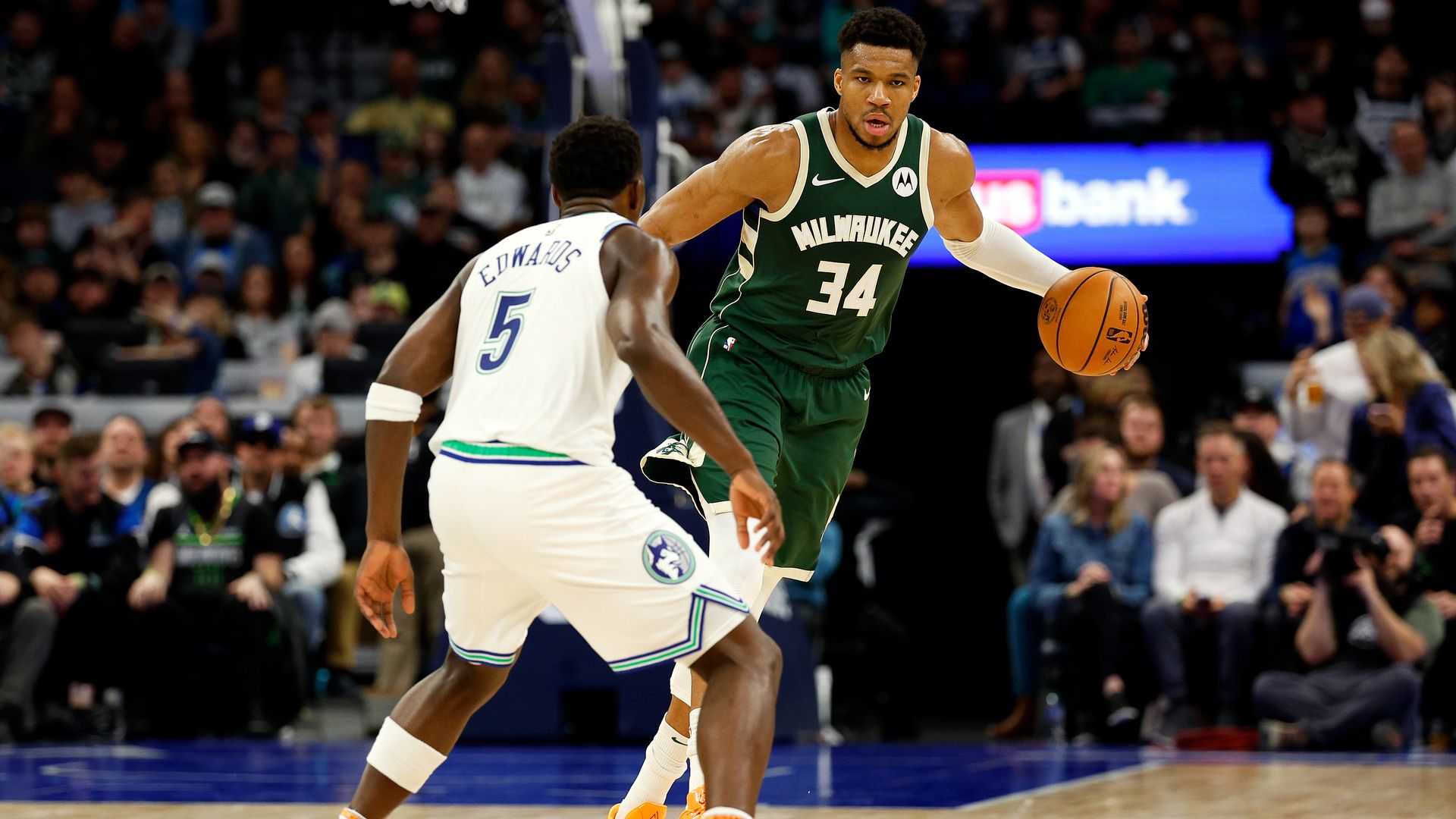 A photo showing Anthony Edwards in a white #5 jersey guarding Giannis Antetokounmpo who is dribbling the ball and wearing a green #34 jersey. 